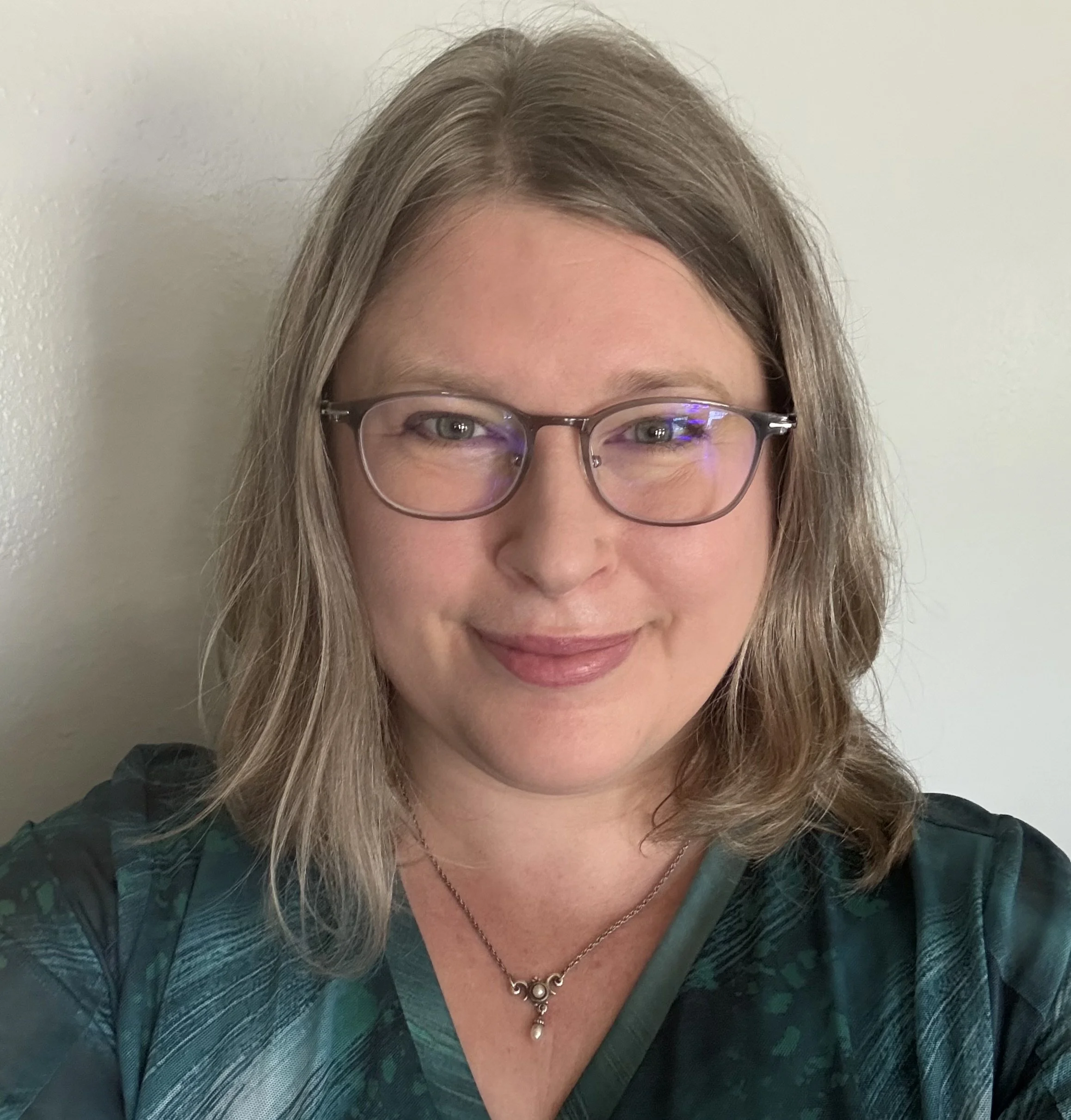 A woman with light brown hair, wearing glasses and a dark green and black patterned top, displaying a necklace with small, ornate charms, smiling softly against a plain white wall.