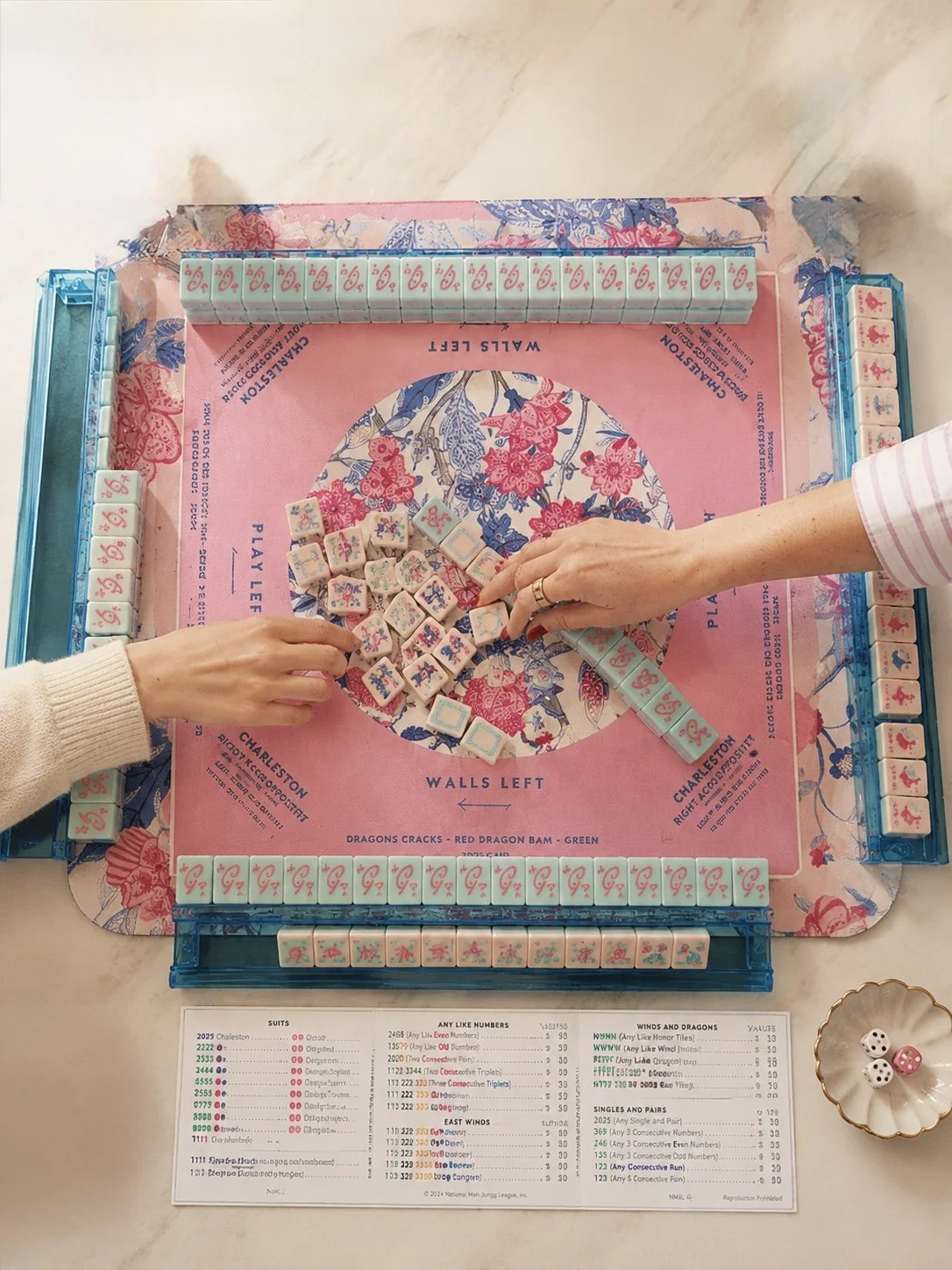 Two people playing Mahjong at a table with a pink floral-patterned mat, with Mahjong tiles and dice on the table.