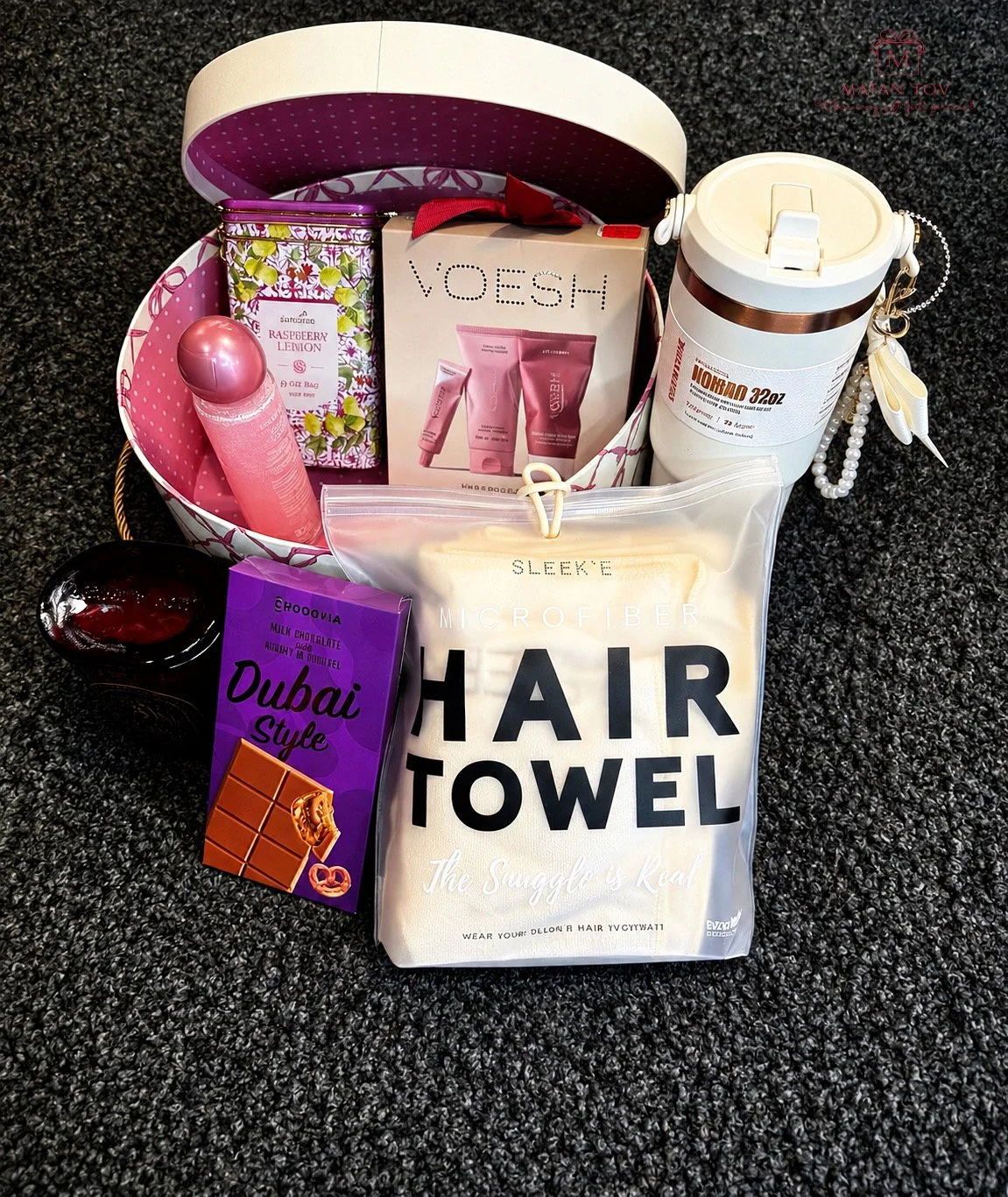 A gift basket containing various personal care and snack items, including a pink spray bottle, a raspberry infusion tea box, a HOODOO Dubai Style chocolate bar, a hair towel, a VOESH skincare set, a white insulated water bottle with a chain, and a pi