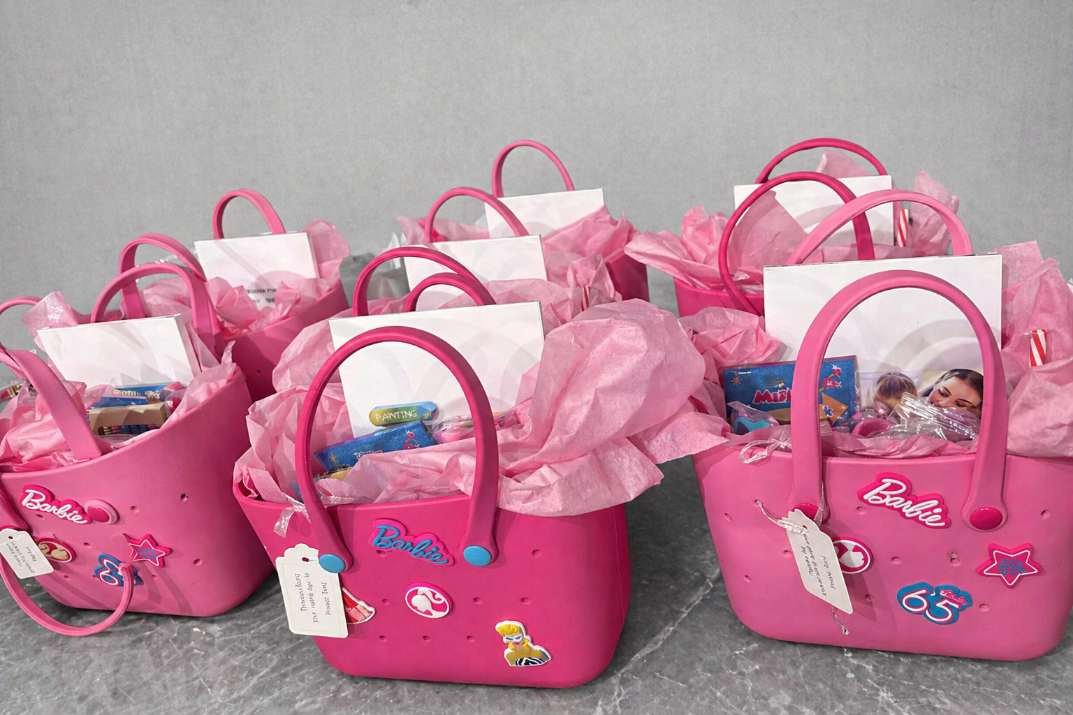 Multiple pink Barbie-themed gift bags filled with pink tissue paper and assorted small toys or gifts, decorated with stickers featuring Barbie and other symbols.
