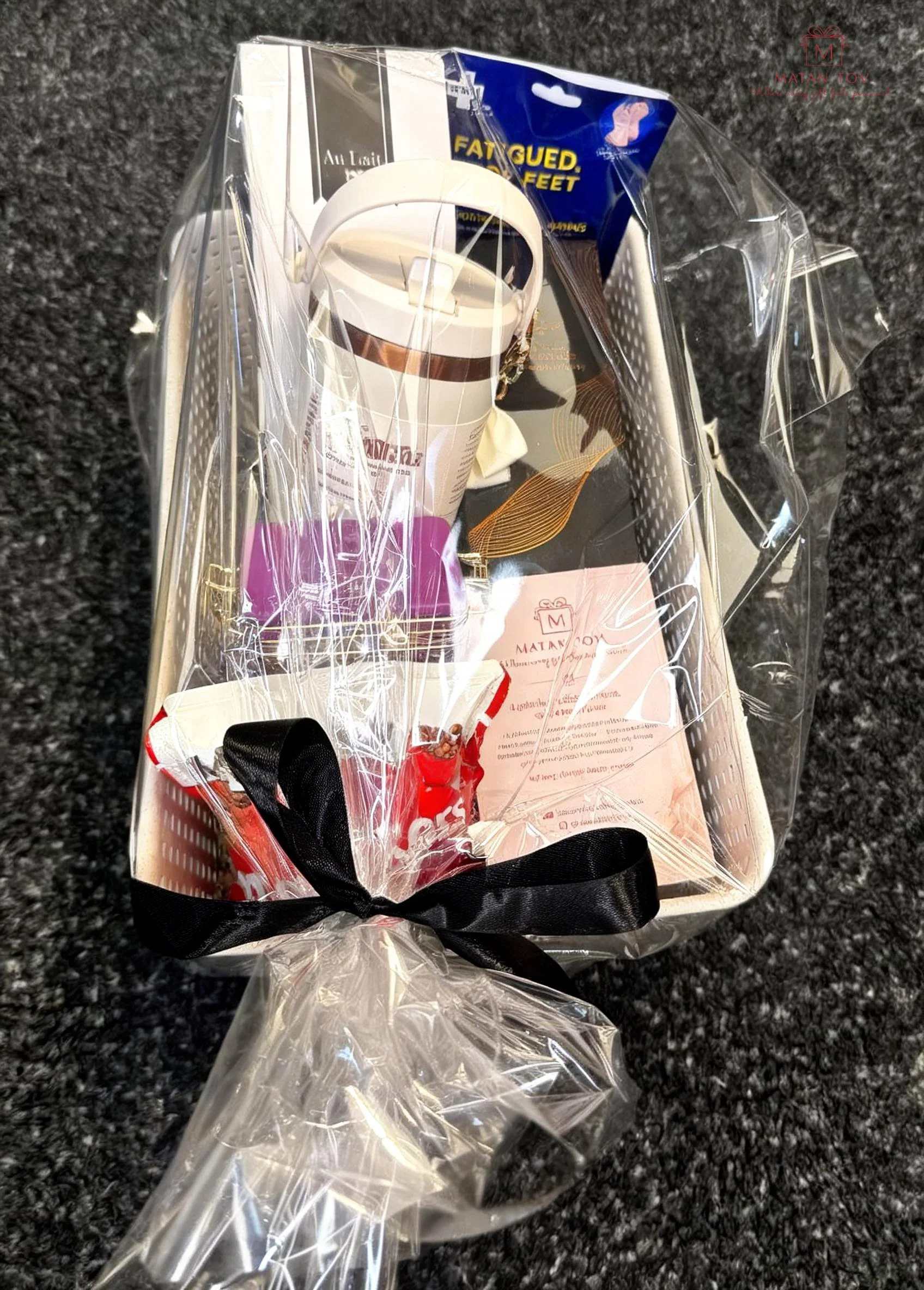 Clear gift bag with a black ribbon, containing a coffee cup, a pair of sunglasses, and brochures or flyers, placed on a dark textured surface.