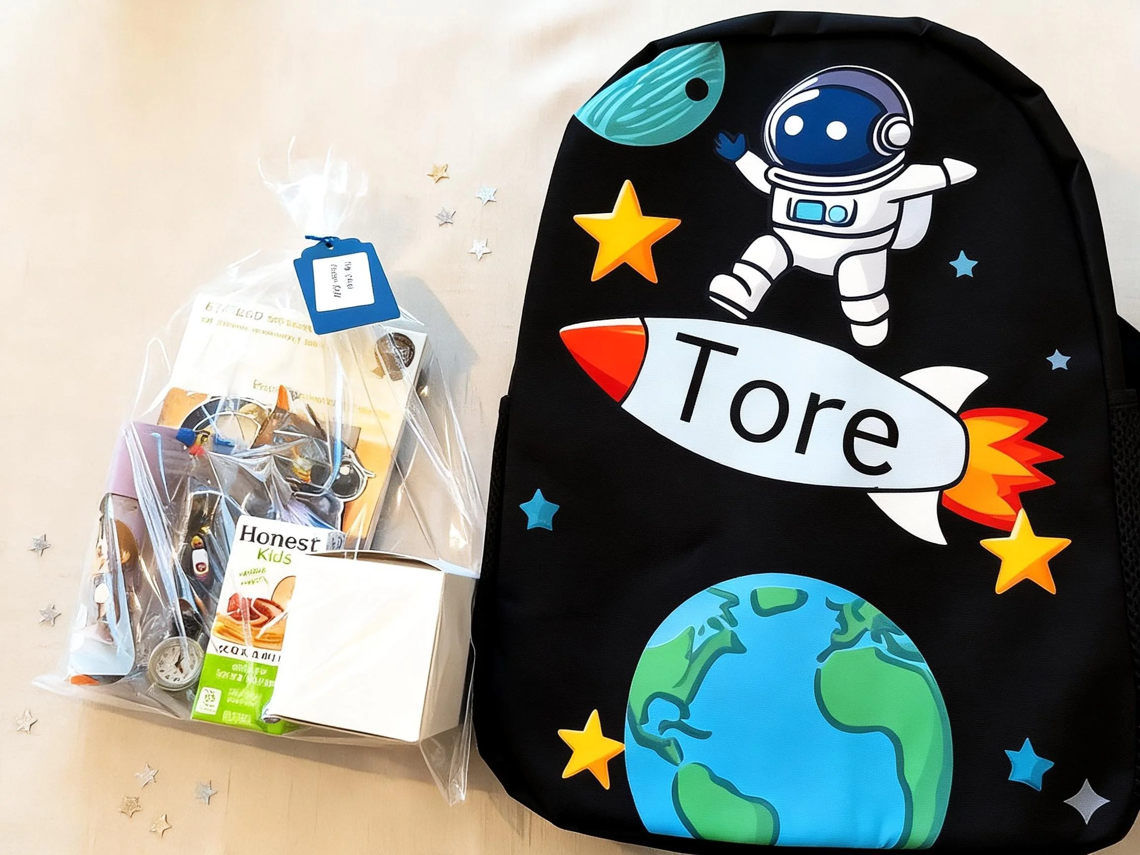 Black backpack with space-themed cartoon design featuring an astronaut and planets, next to a clear plastic bag of assorted candies on a white surface decorated with small silver and blue star confetti.