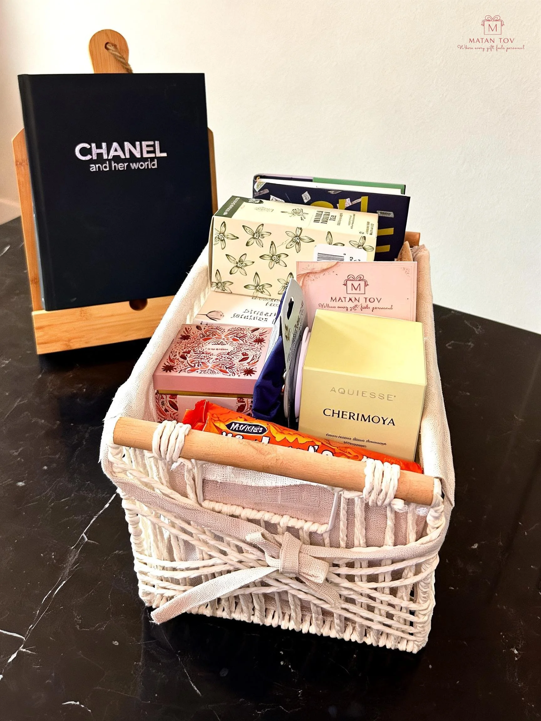 A white woven basket filled with various small gift boxes and chocolates on a black marble surface, with a black book titled 'Chanel and her world' on a small wooden stand behind it.
