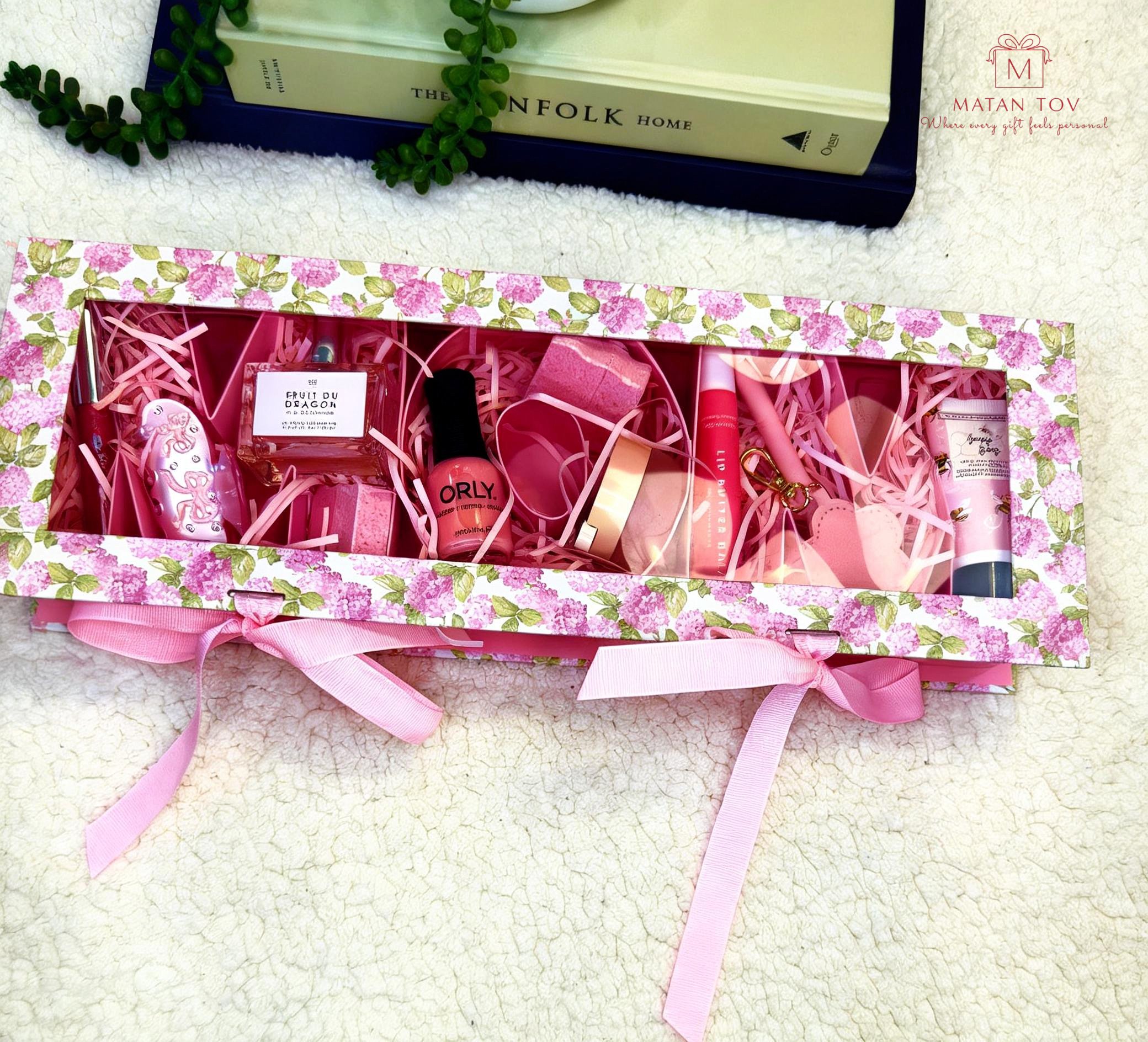 Pink gift box with pink ribbon filled with pink and purple beauty products, placed on a fluffy cream-colored surface.