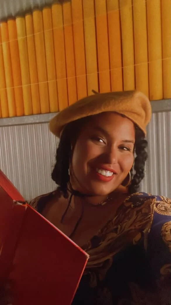 A woman smiling with a beret, dark curly hair, gold hoop earrings, in front of a background with yellow and gray paneling, holding a red box.