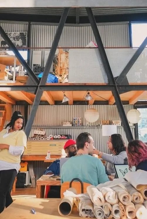 A group of people sitting at a wooden table in a modern industrial-style cafe, with people chatting and a woman standing nearby. The cafe has a high ceiling, large window, and industrial decor with rolls of paper or fabric on the table.