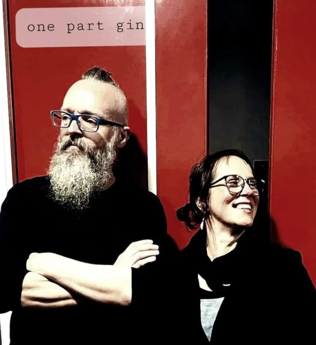 A man with a beard, glasses, and a distinctive hairstyle with the sides shaved and a top knot, standing with his arms crossed. A woman with glasses and short dark hair is smiling beside him. The background is a red wall with a sign that reads "one pa