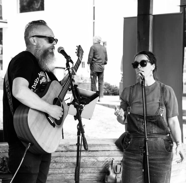 A man with a beard and sunglasses playing acoustic guitar and singing into a microphone, while a woman with sunglasses and overalls sings into another microphone, outdoors in front of a modern building.