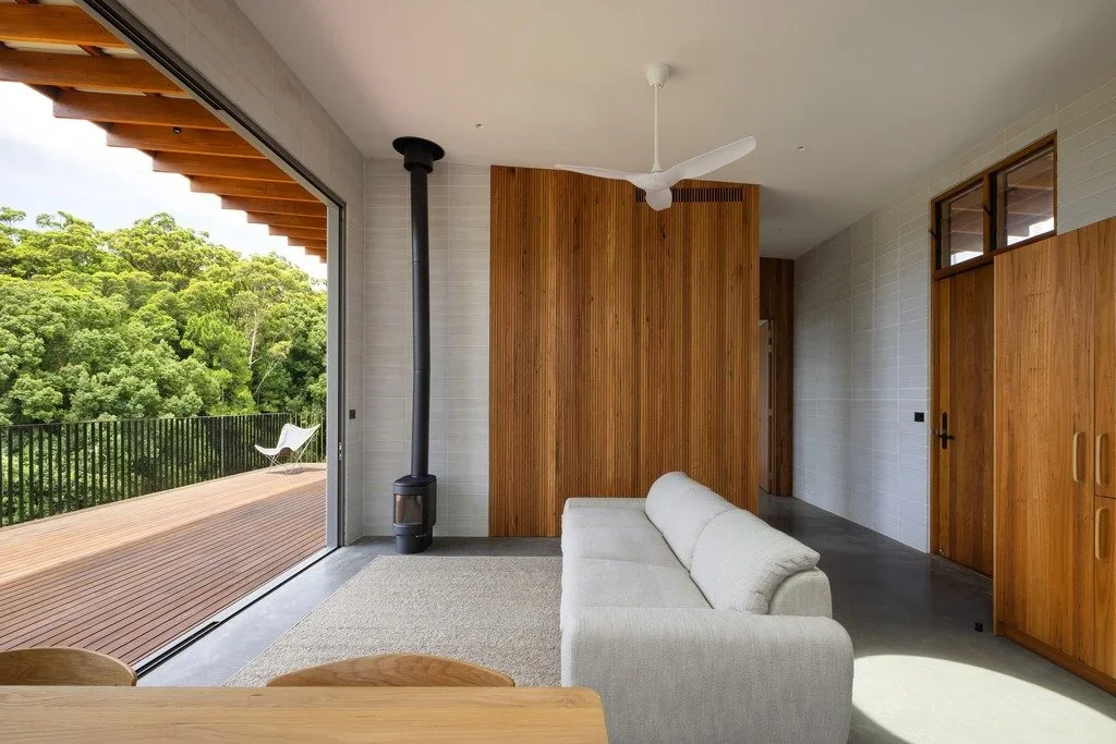 Living room with white sofa, wooden accent wall, large sliding glass door leading to a deck with outdoor seating, and green trees in the background.
