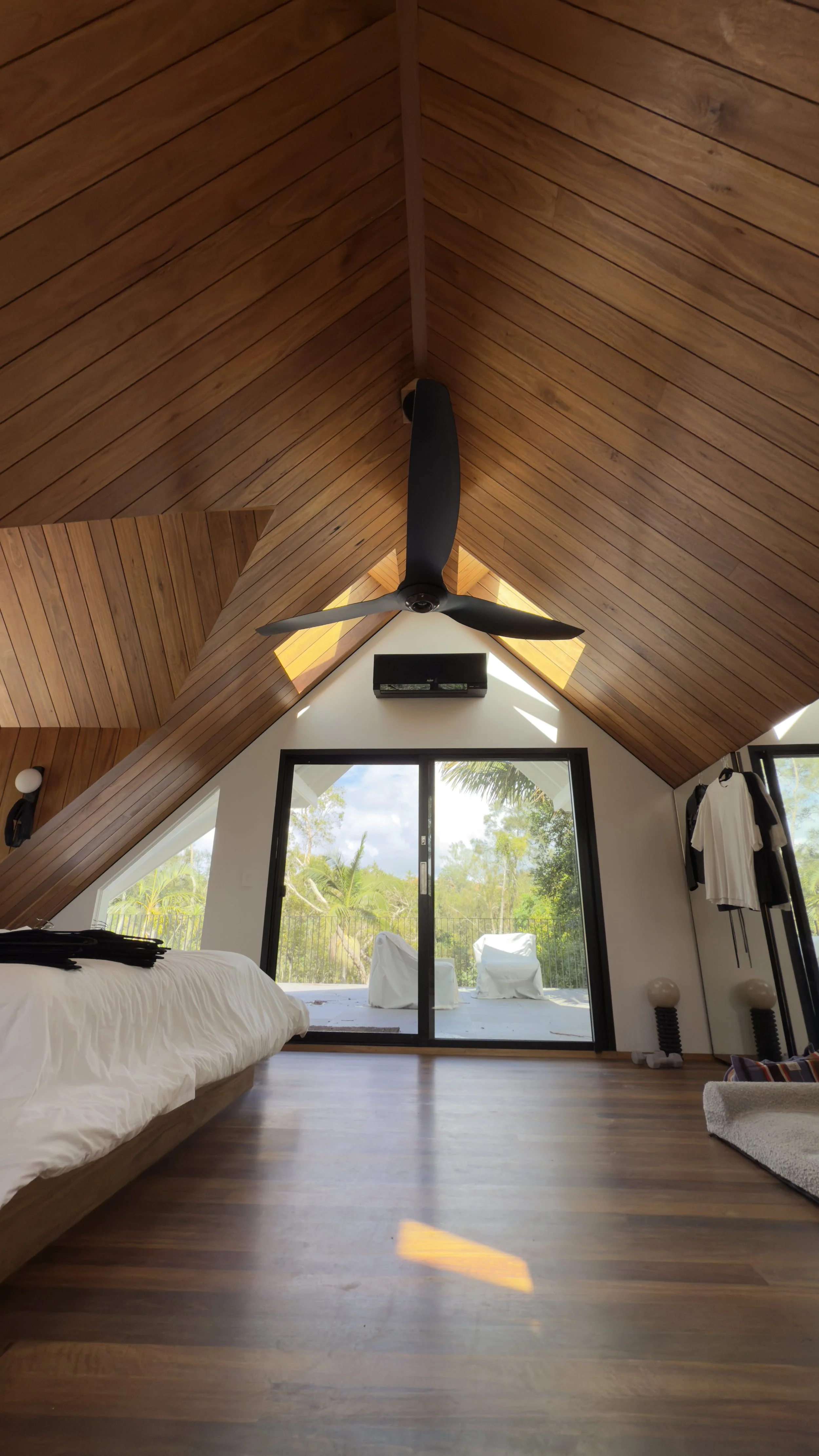 Interior of a bedroom with a vaulted wooden ceiling, ceiling fan, large sliding glass door opening to a balcony, and natural light.