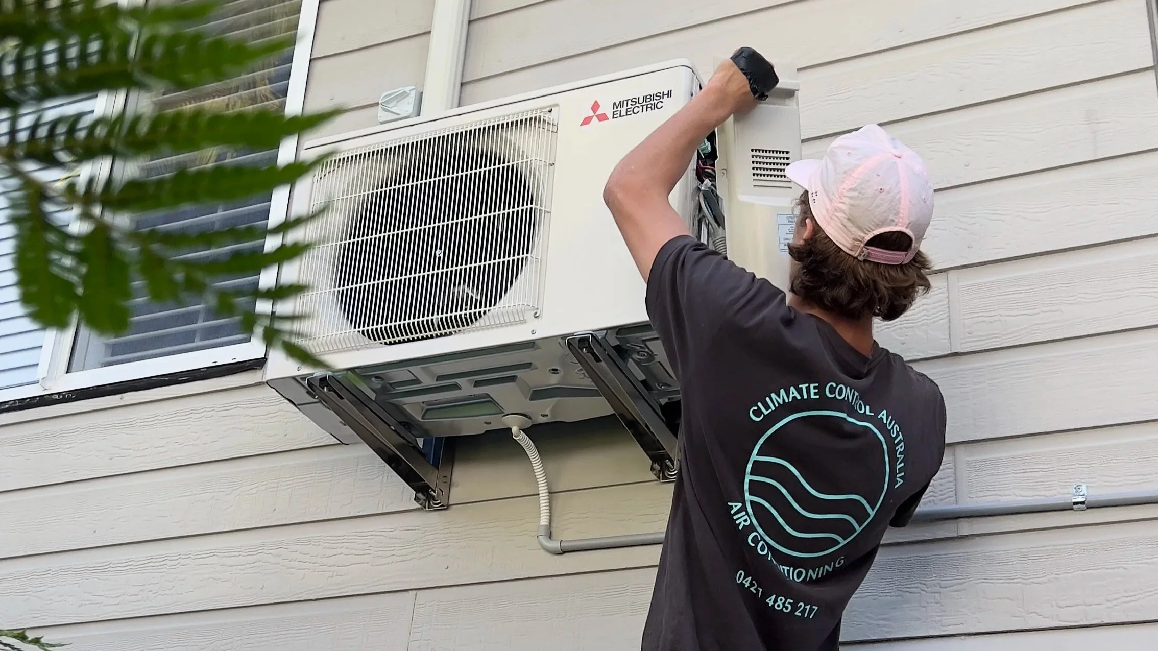 A person wearing a black T-shirt and a pink baseball cap installing or repairing a Mitsubishi Electric air conditioning unit outside a house with beige siding.