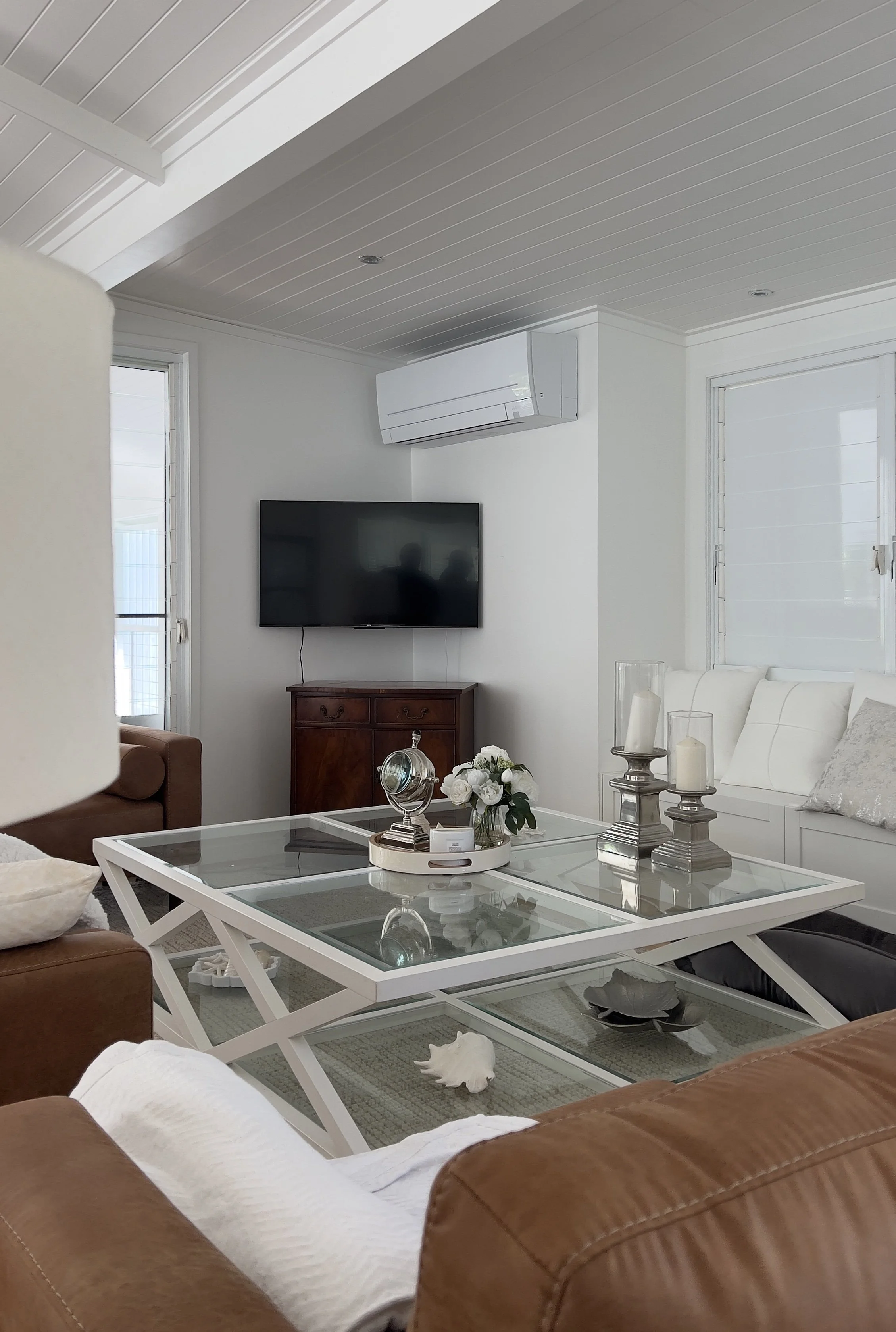 Living room with white walls and ceiling, wall-mounted TV, wooden cabinet, white couch, glass coffee table with decorative items, candles, and a white pillow, natural light from windows.
