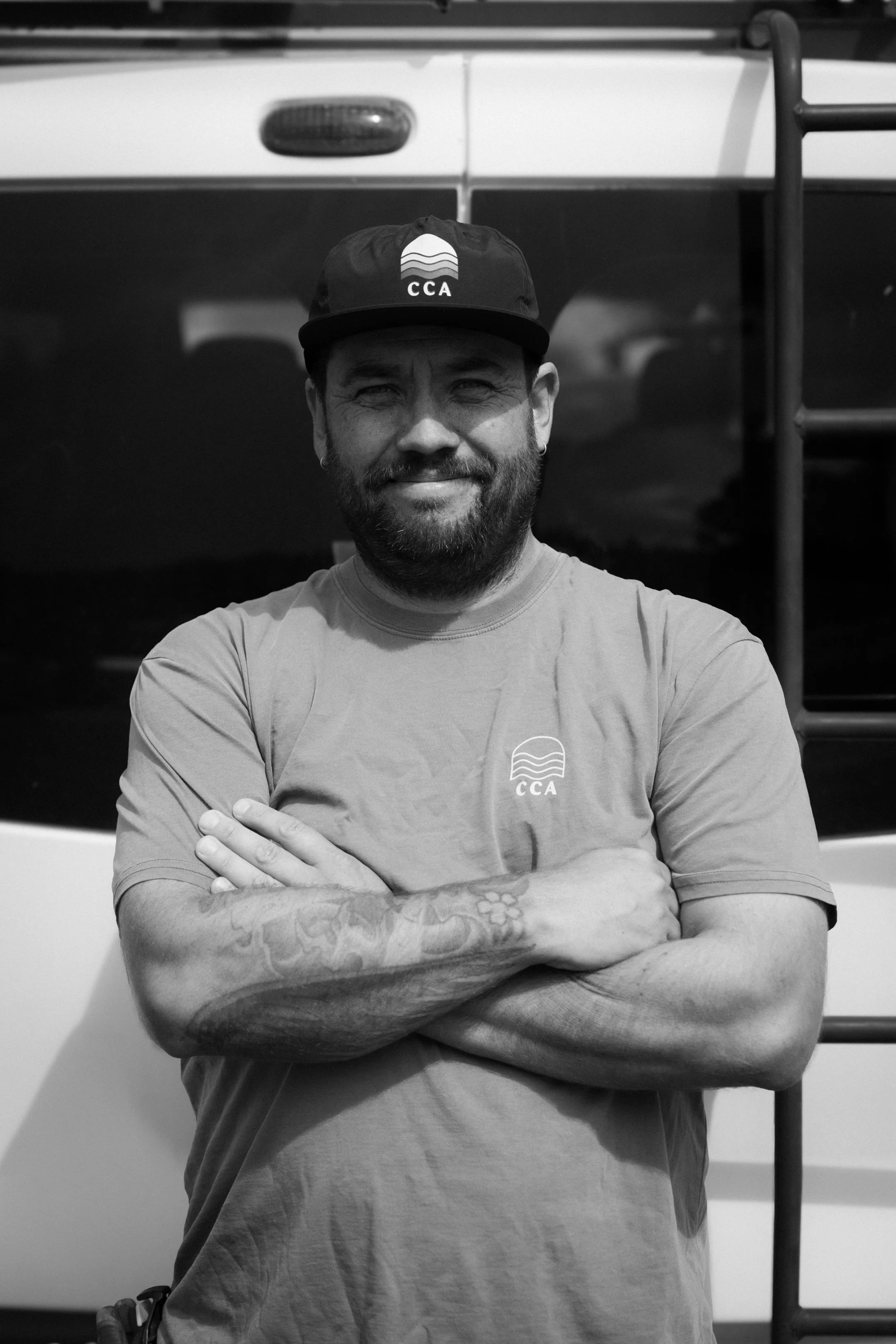 A man with a beard and tattoos on one arm, wearing a cap and t-shirt with the logo 'CCA', standing with arms crossed in front of a vehicle with ladders.