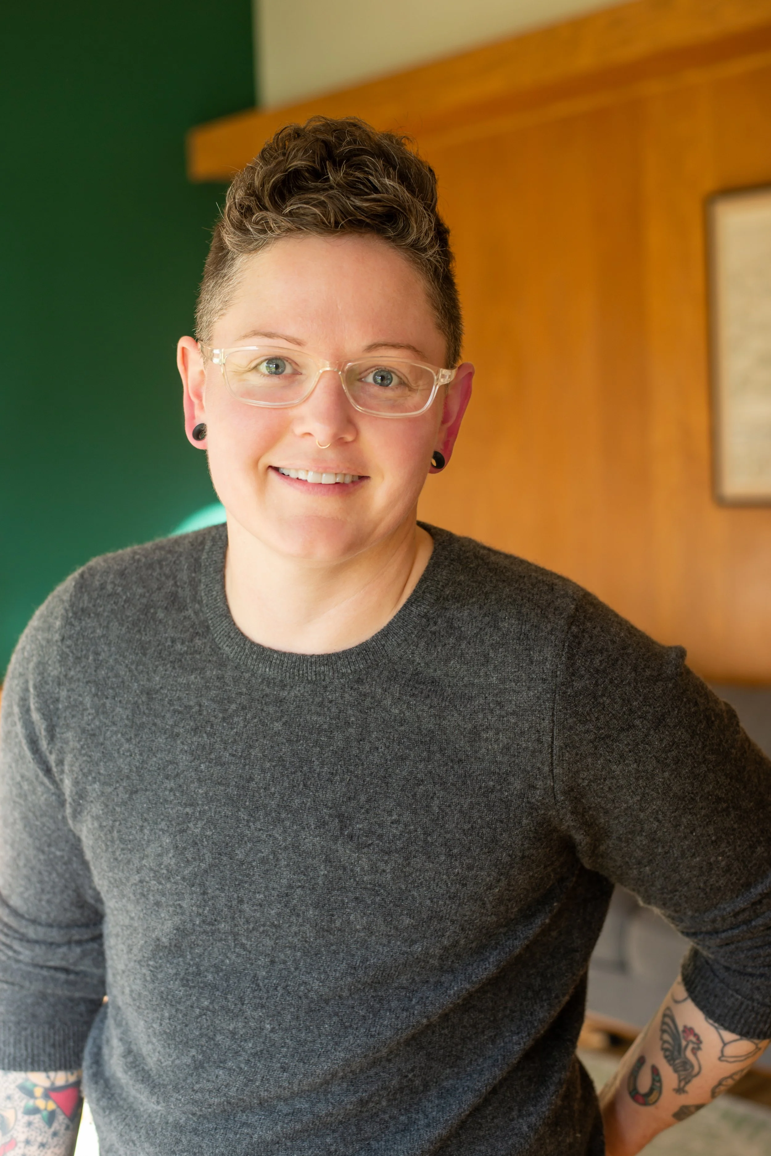A woman with short, curly hair and glasses, smiling, wearing a dark sweater, with visible tattoos on her arm.