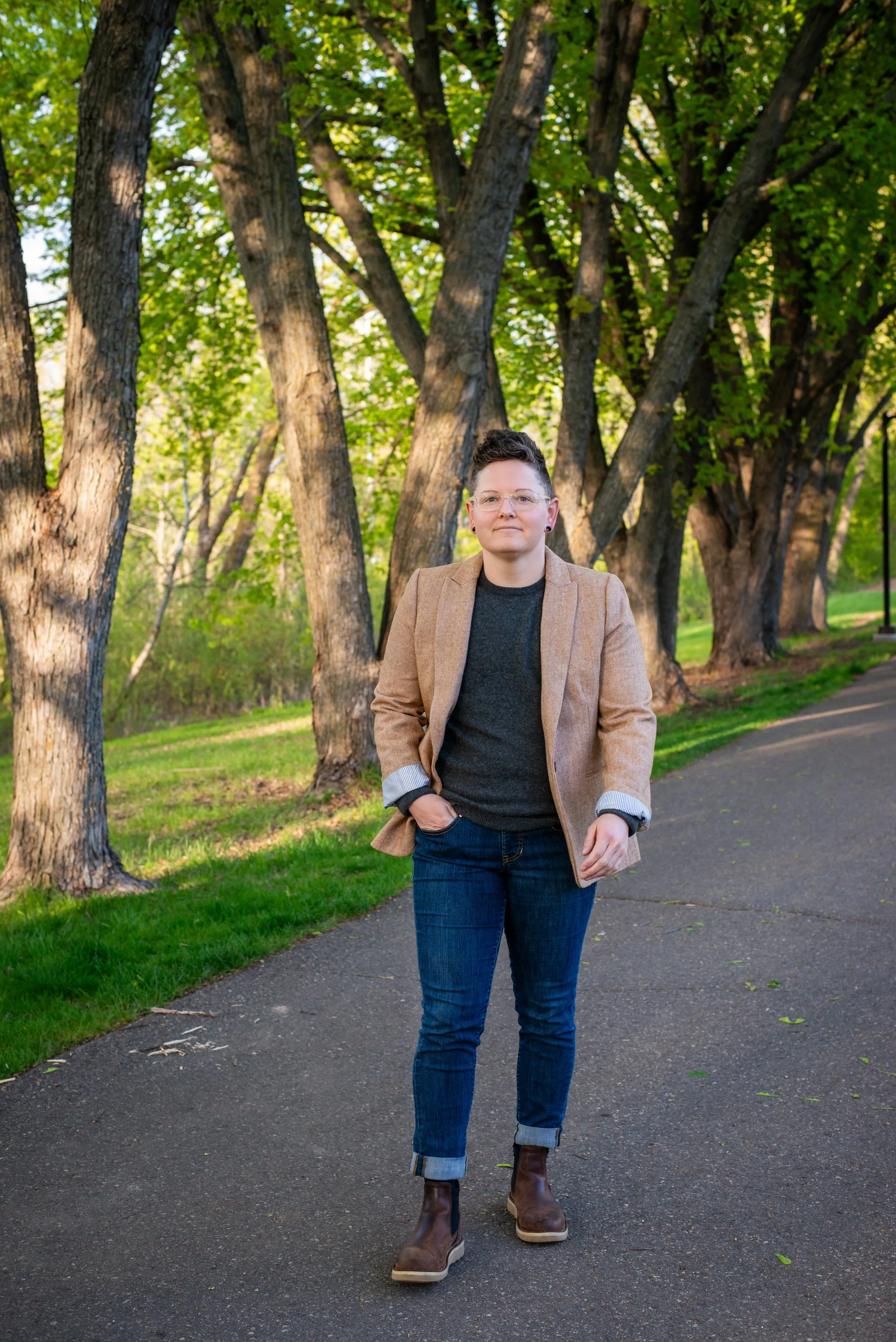 A woman with short curly brown hair and glasses, dressed in a light brown blazer, dark sweater, and jeans, walking on a paved path through a park with large trees and green grass.