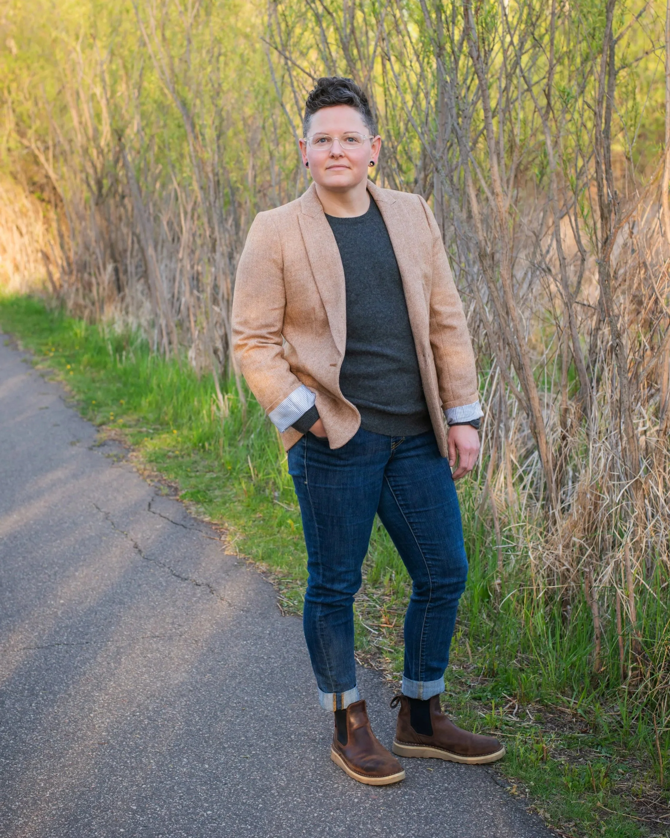 A woman with short curly brown hair and glasses, standing on a paved path beside a row of tall grass, wearing a light brown blazer, dark sweater, jeans, and brown boots, with her hands in her pockets.