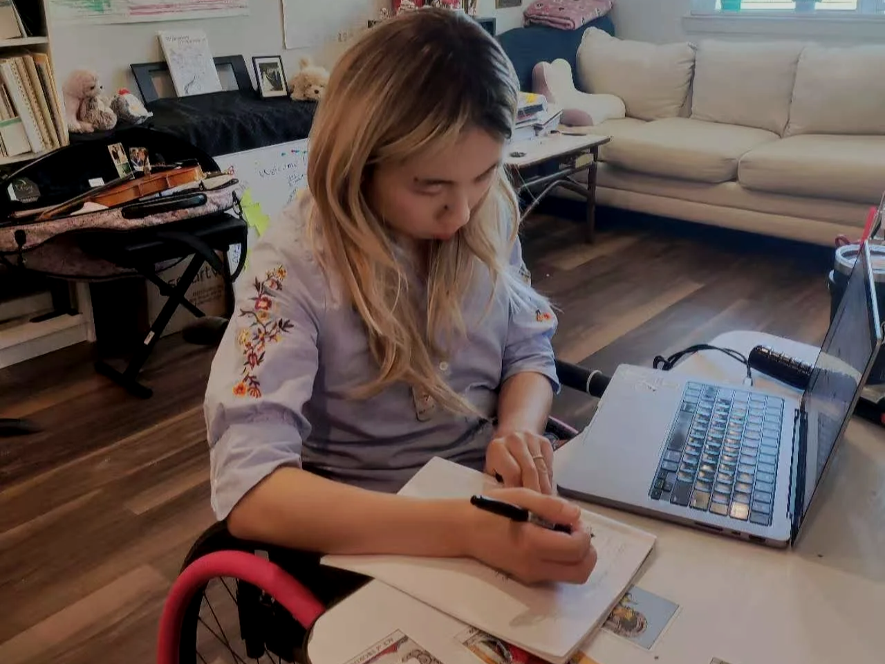 A woman with long blonde hair is sitting at a table in a living room, writing in a notebook with a pen. She is wearing a light-colored shirt with embroidered floral patterns on the sleeves. A laptop is open on the table next to her. The background includes a beige couch, a table with books, stuffed animals, and picture frames.