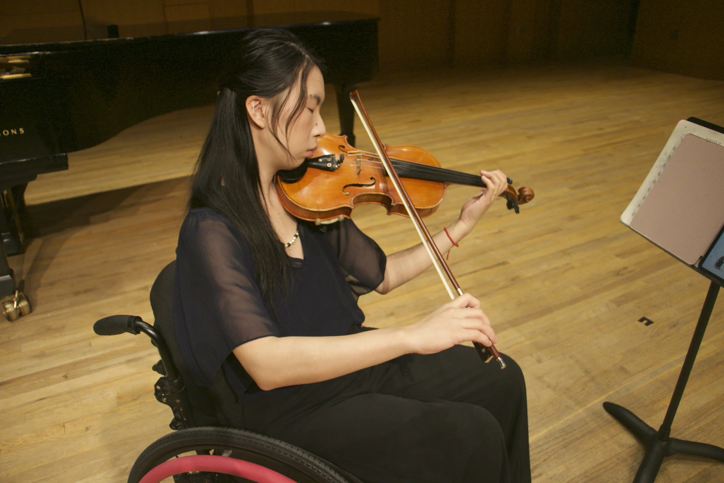 A woman playing the violin while seated in a wheelchair in a music room with wooden flooring.