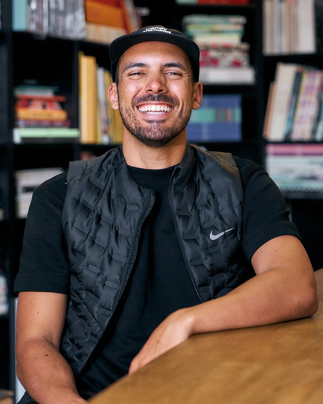A smiling man wearing a black cap, black Nike vest, and black t-shirt, sitting at a wooden table with a background of bookshelves filled with books.