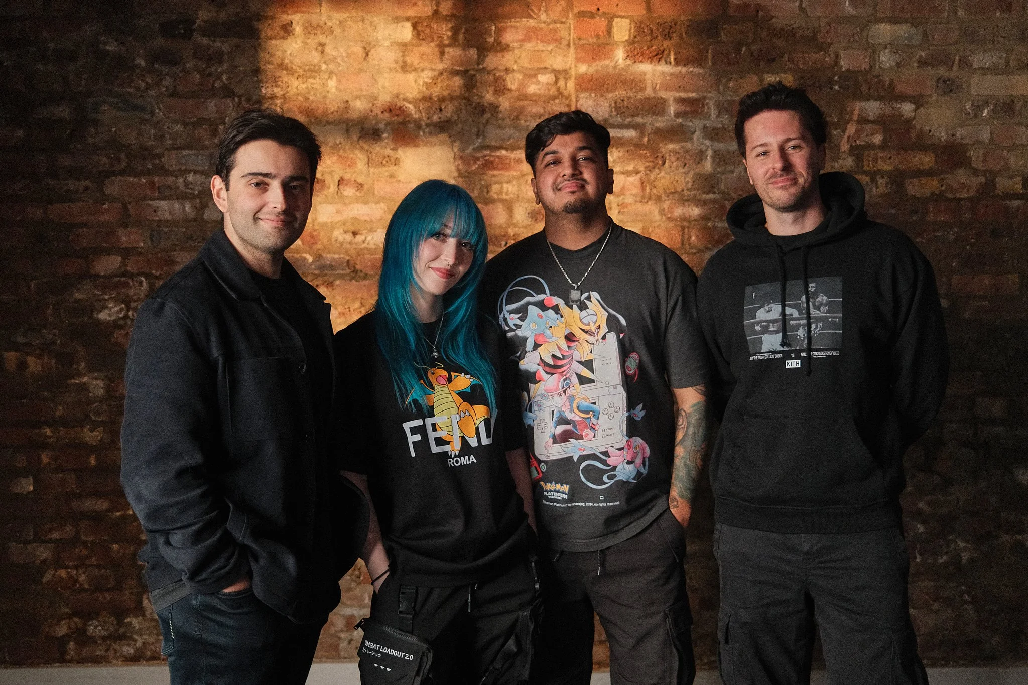 Four young adults standing together in front of a brick wall, smiling for the camera. The group includes two men and one woman with blue hair, all dressed casually.