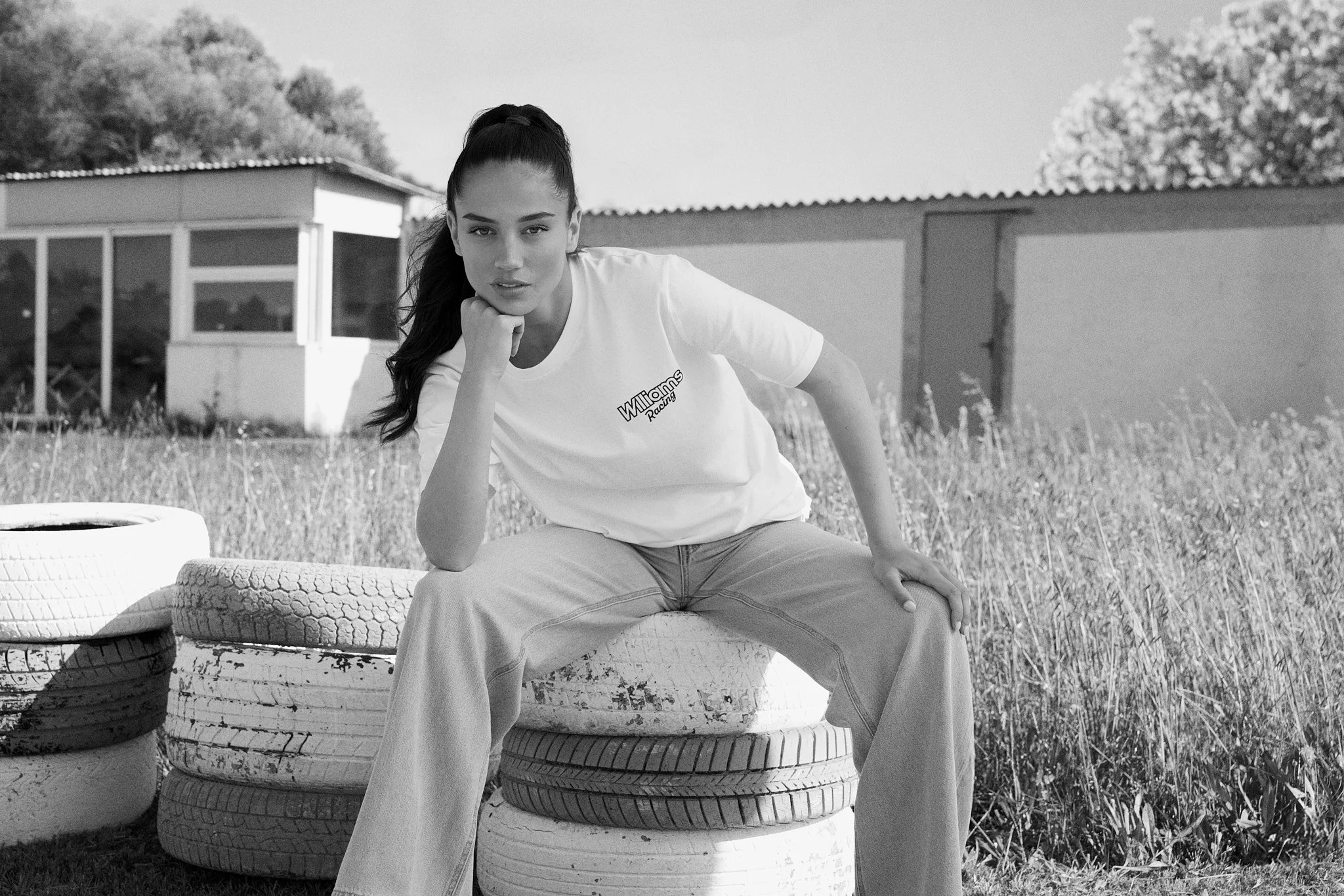 A woman with long dark hair tied in a ponytail, wearing a t-shirt that says 'Willoms Racing,' sitting on stacked tires outdoors. She is resting her chin on her hand and looking at the camera. The background features a building and some trees.
