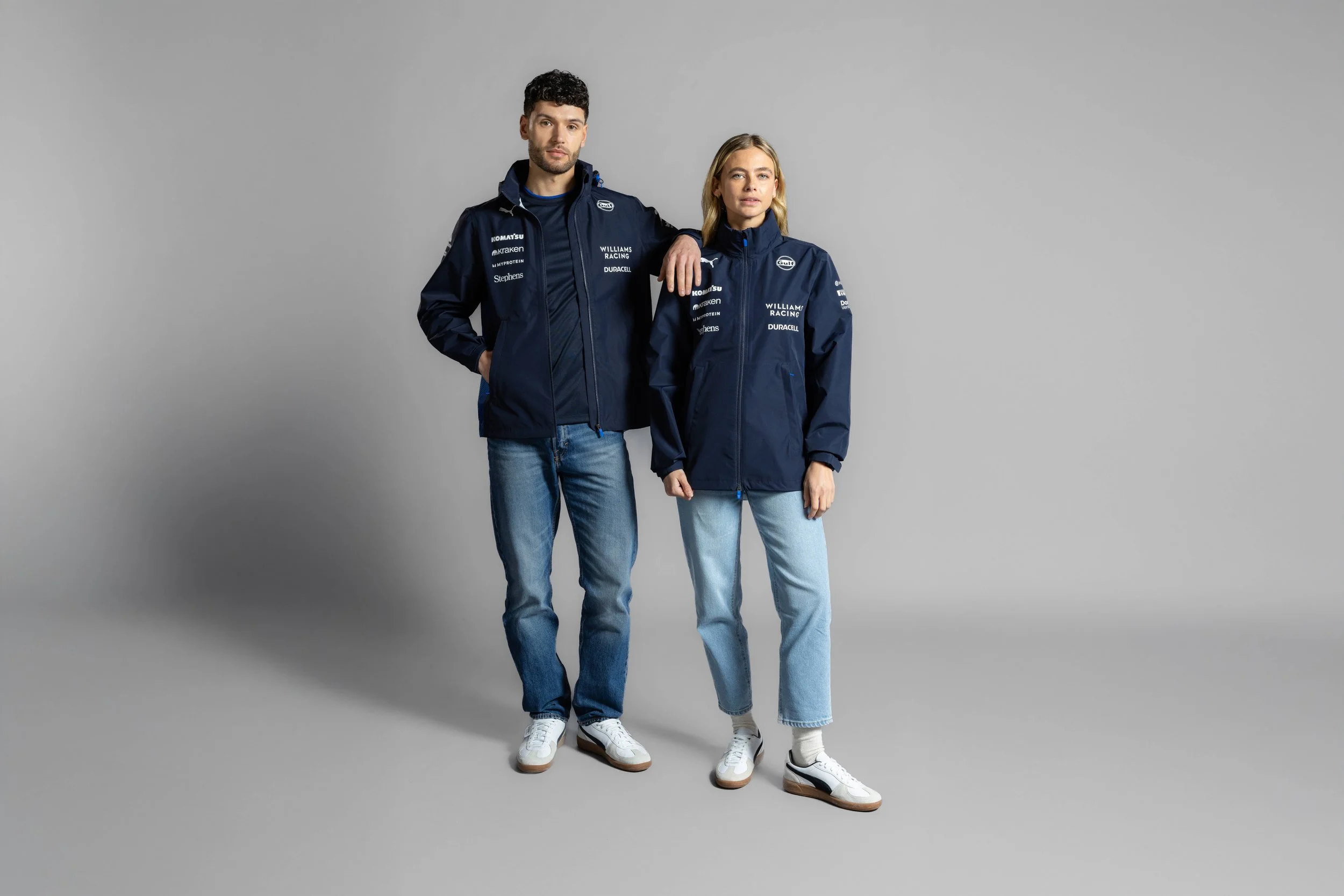 A man and a woman standing side by side against a plain gray background, wearing navy blue racing jackets with sponsor logos, jeans, and white sneakers.