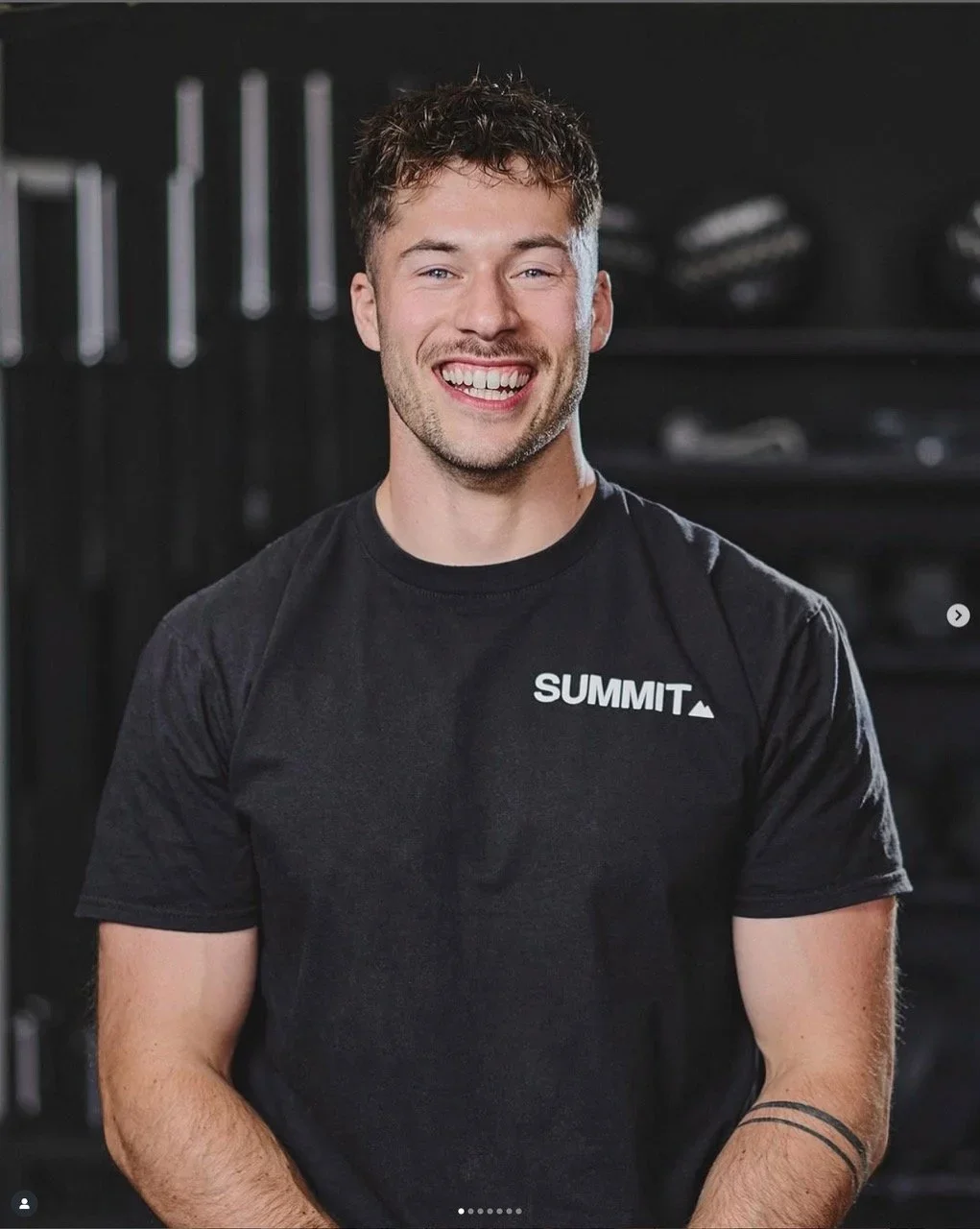 Young man smiling in a gym, wearing a black T-shirt with 'SUMMIT' logo, with gym equipment in the background.