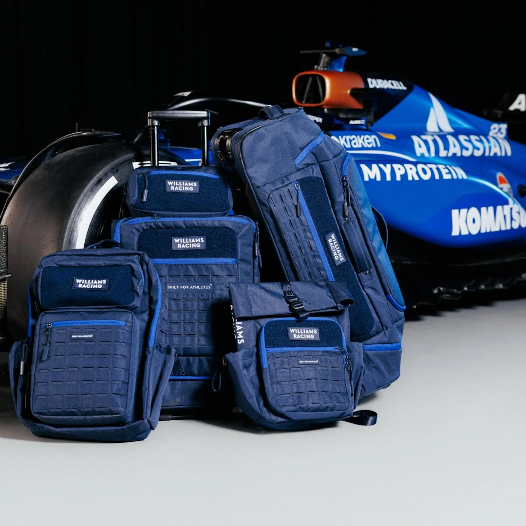 Collection of blue Williams Racing team bags and backpacks arranged next to a blue Formula 1 car with various sponsor logos.