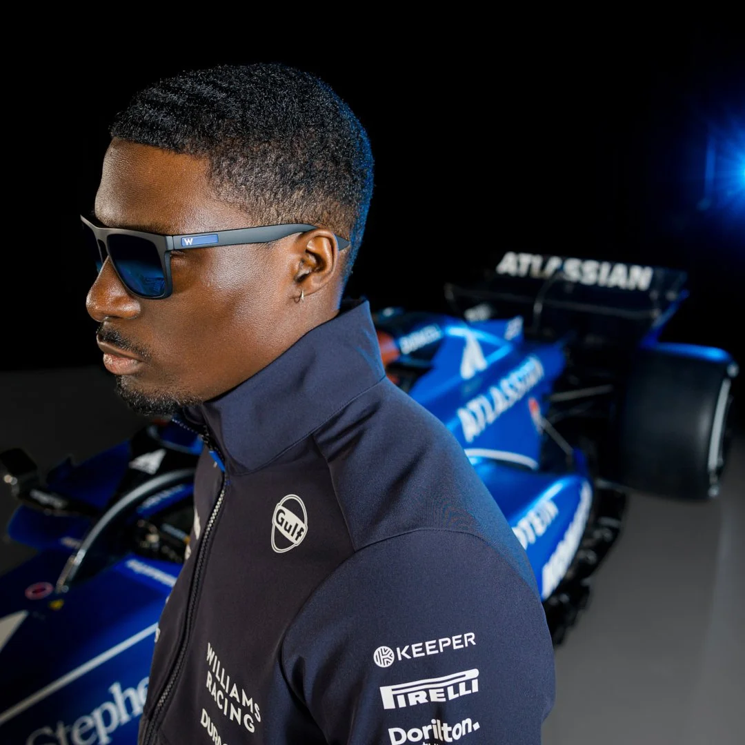 Close-up of a man wearing sunglasses and a racing jacket standing in front of a blue Williams Racing Formula 1 car with sponsors Gulf, Pirelli, and others.