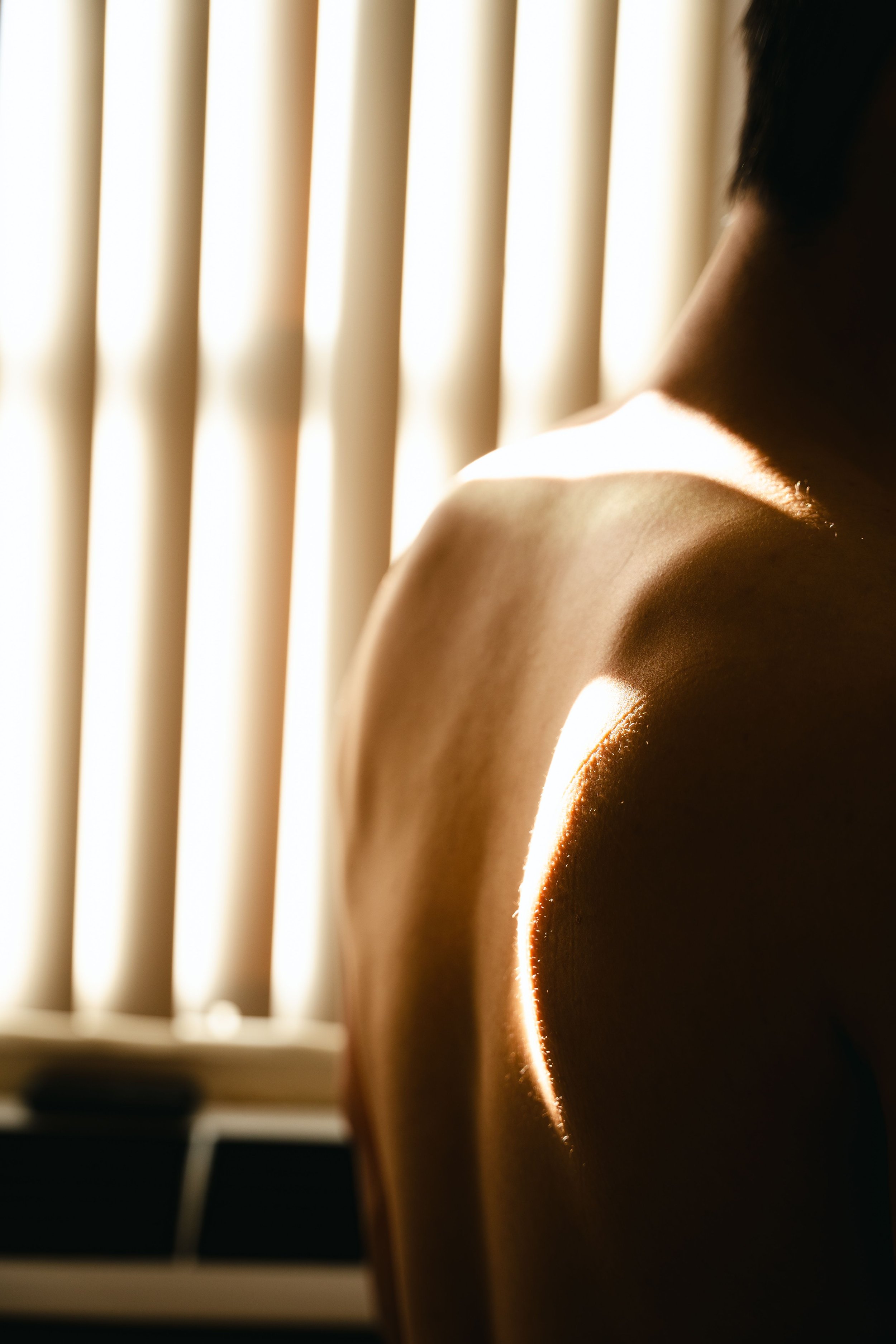 Close-up of a person's shoulder illuminated by sunlight, with a background of window blinds.