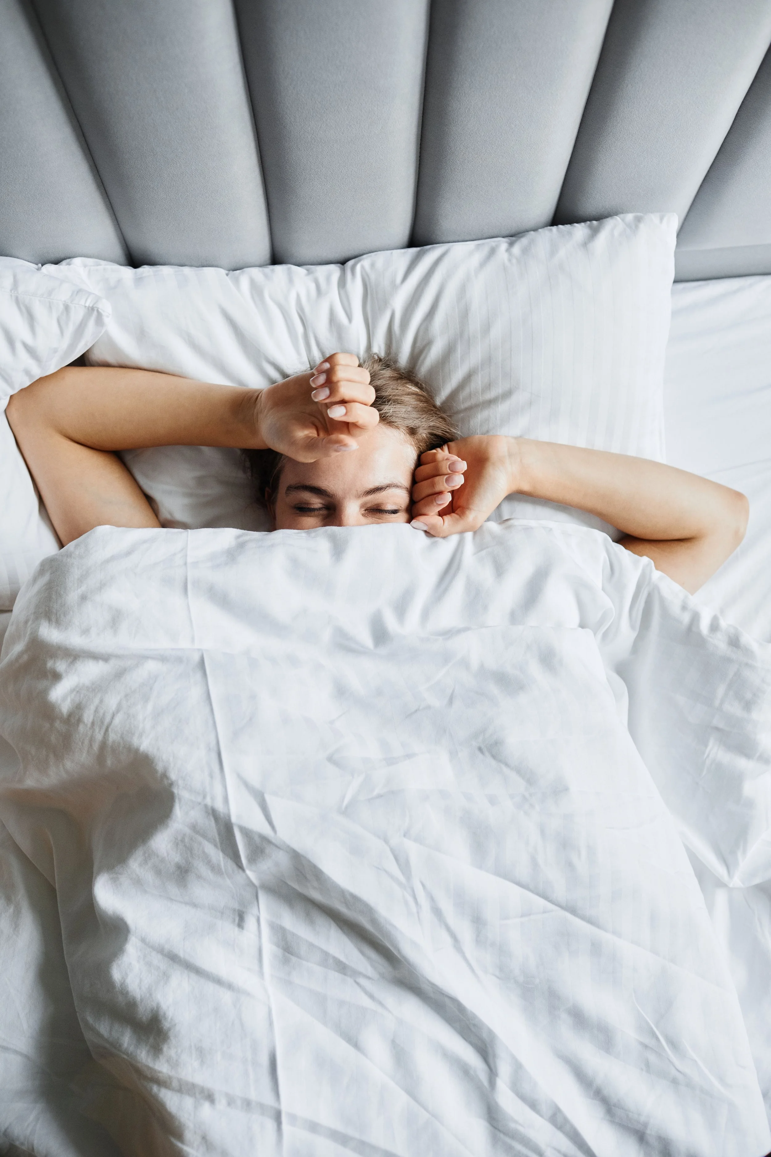 A woman lying in bed with a surprised or shocked expression, covering her face with her hands, partially hidden under white bedding, in a bedroom with a gray upholstered headboard.