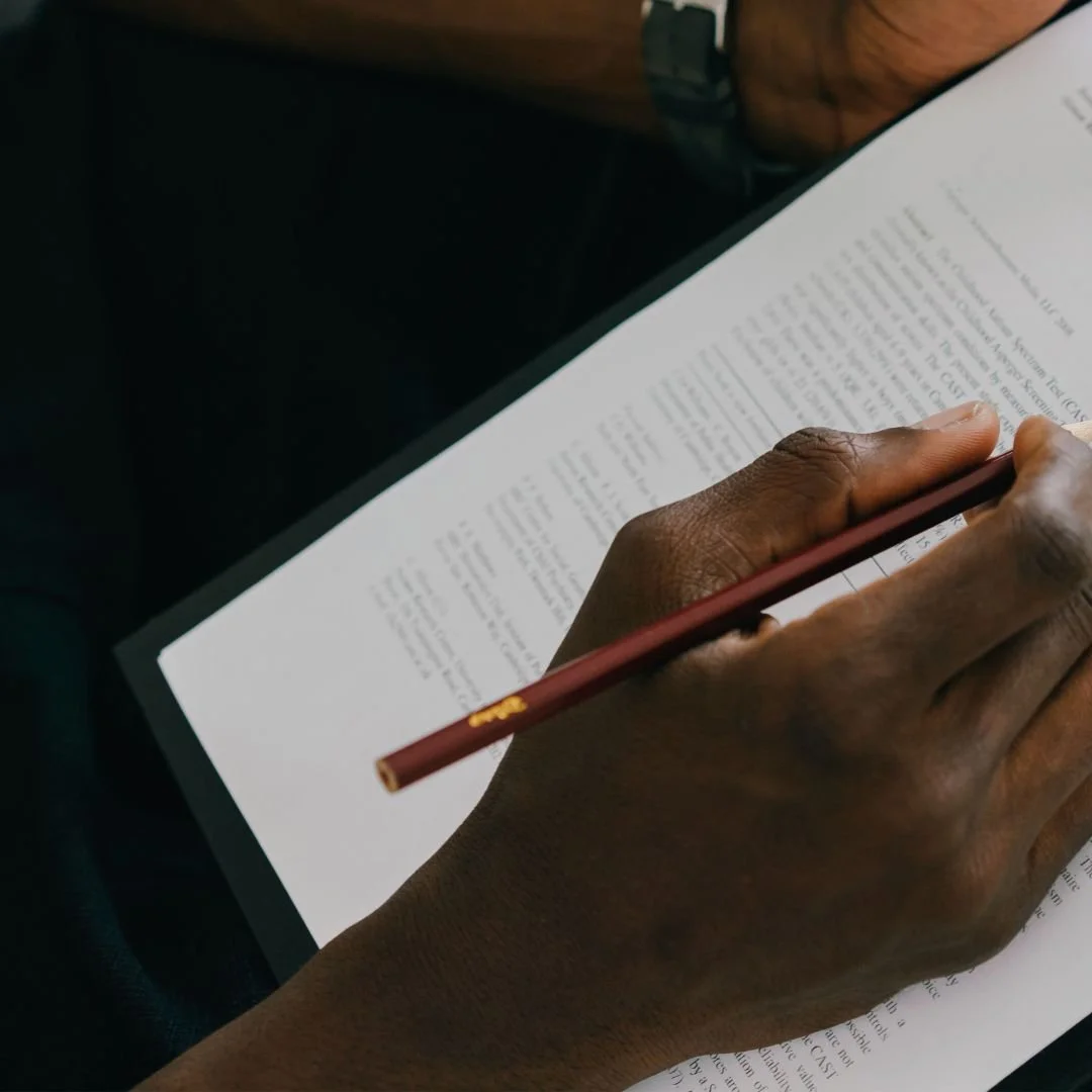 A person with dark skin writing on a paper with a red pen.