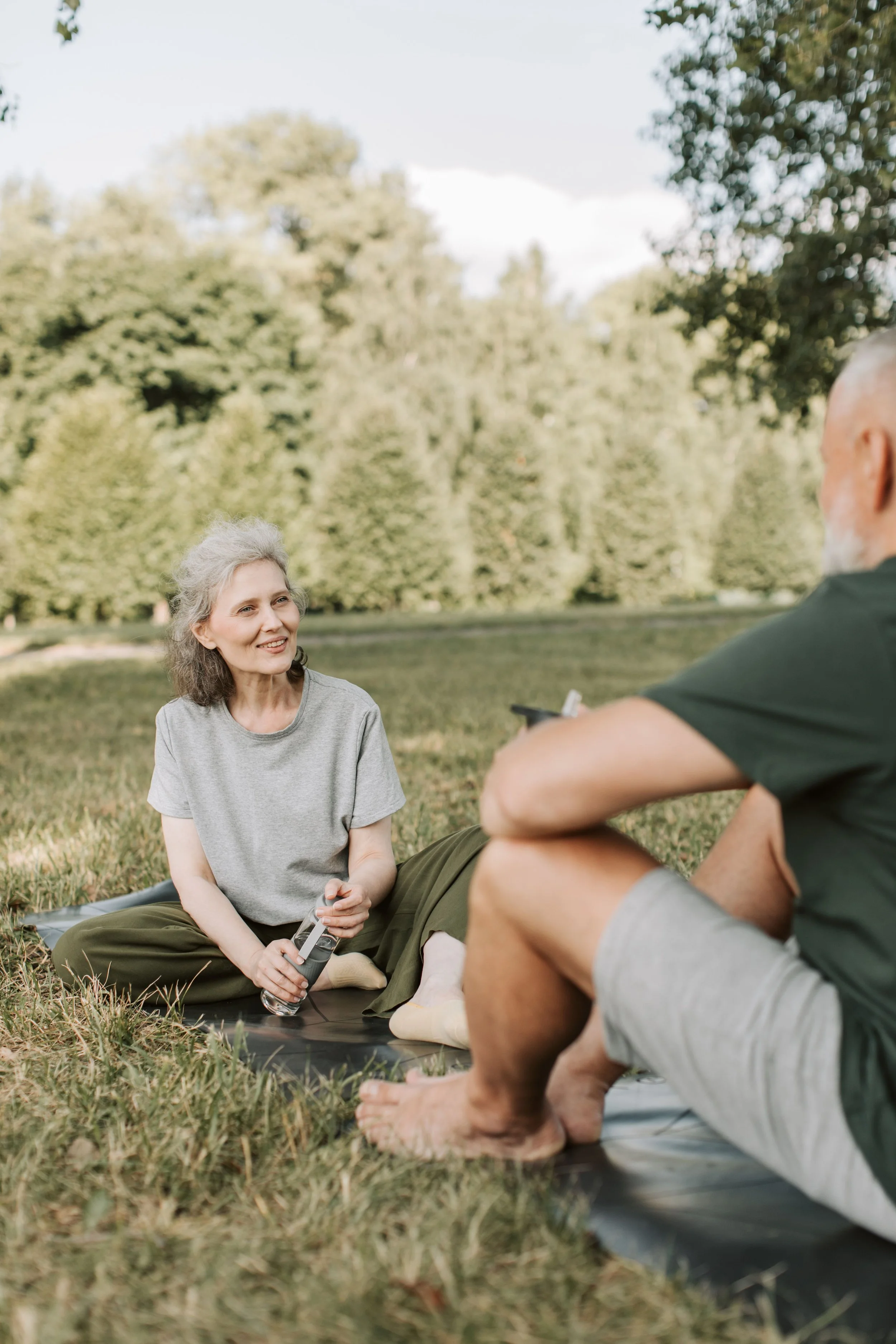 An elderly woman with gray hair sitting outdoors on grass, smiling, holding a water bottle, and engaging in conversation with a man whose face is not visible. They are in a park with trees in the background.