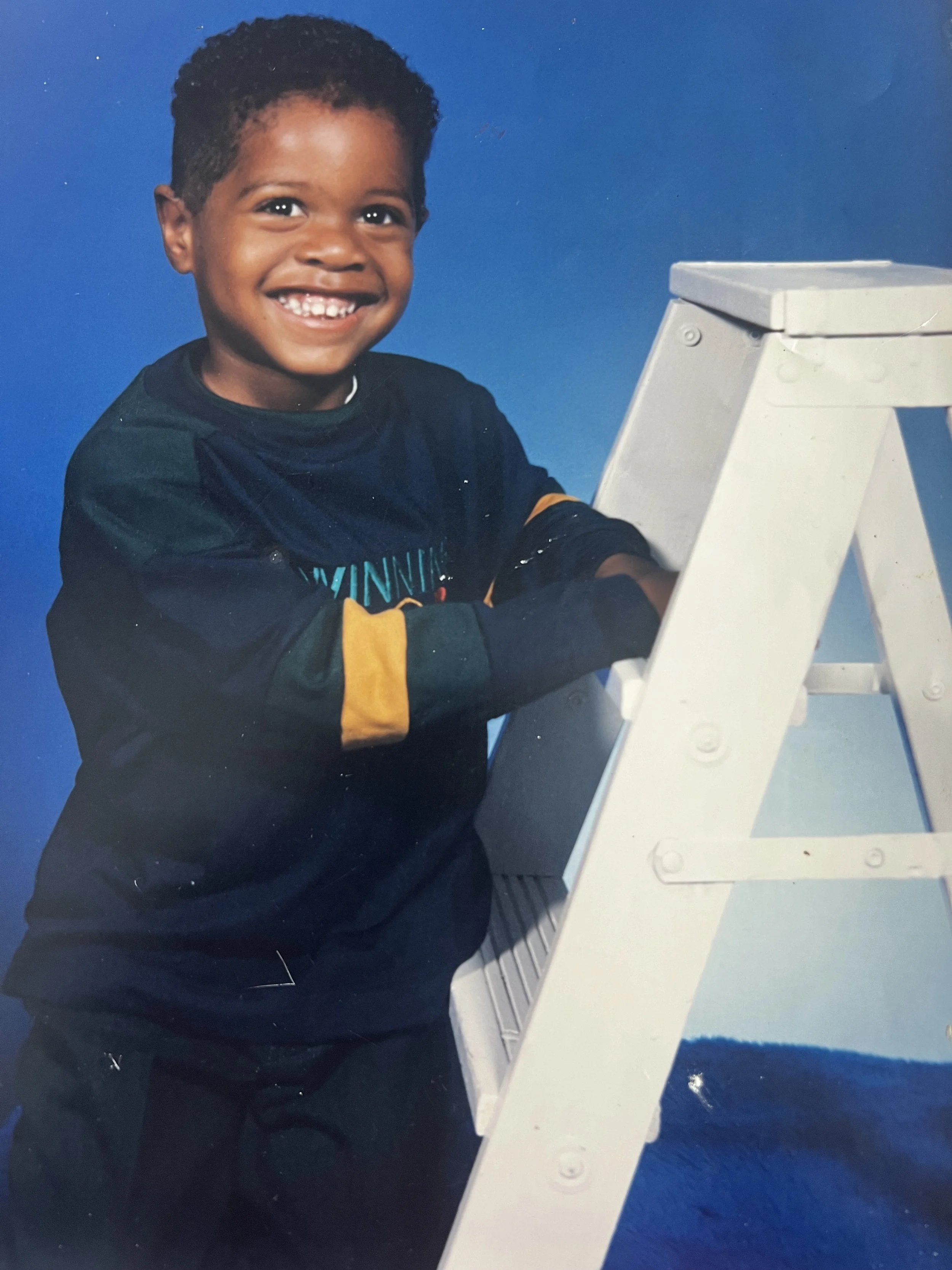 A smiling young boy with short curly hair, wearing a dark long-sleeve shirt with yellow accents, stands next to a white stepladder against a blue background.