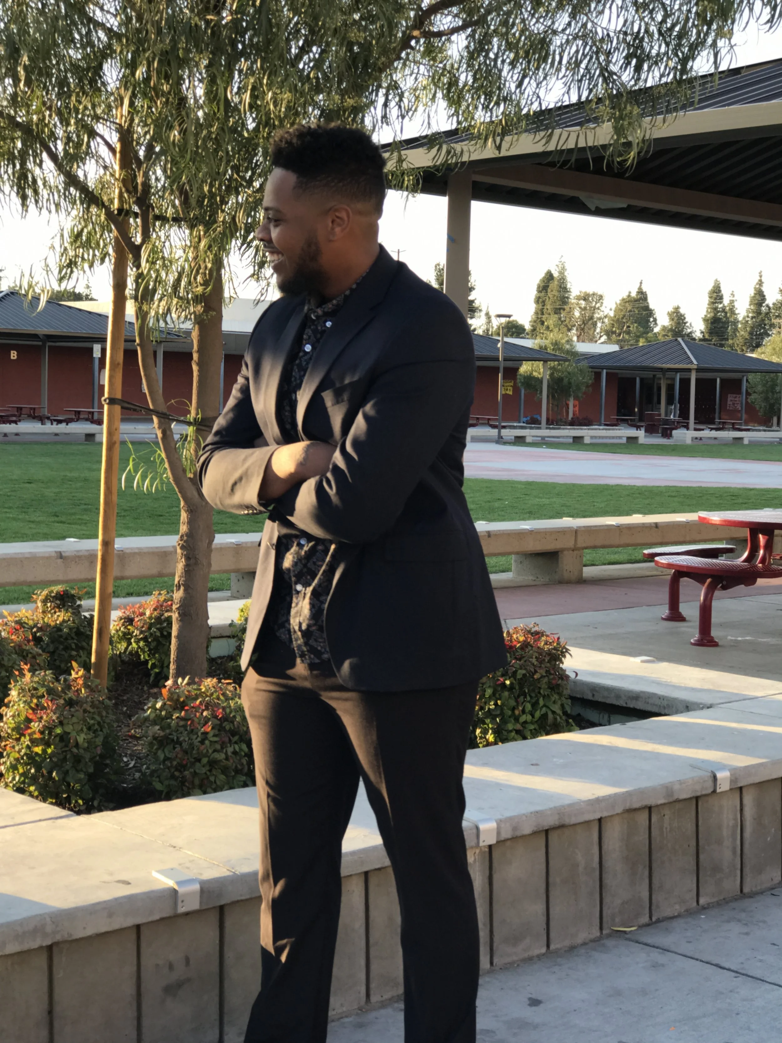 A man with a beard and short hair, dressed in a dark blazer and shirt, smiling with arms crossed, standing outdoors near a planter with flowers and a small tree, with buildings, trees, and a pavilion in the background.