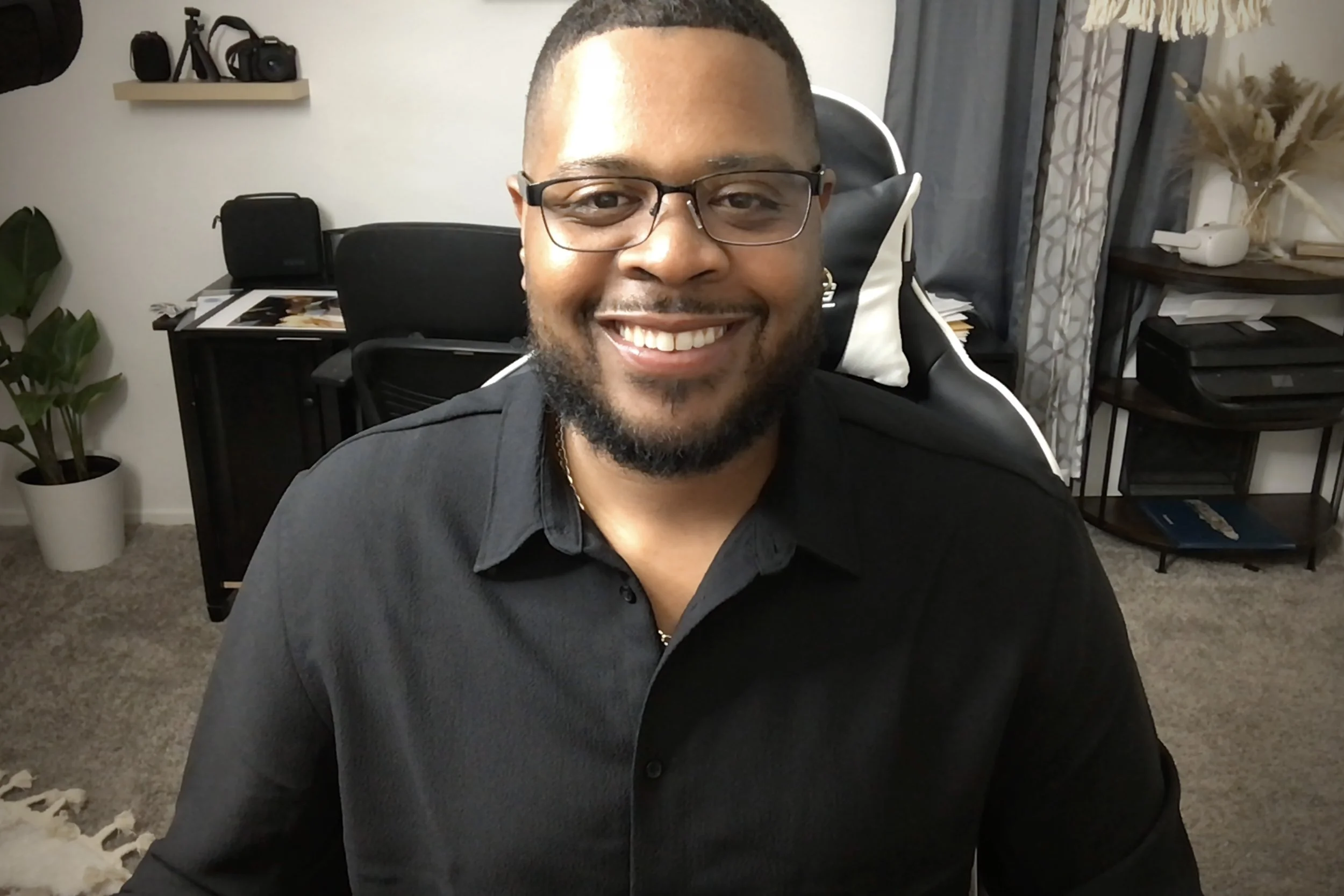A smiling man with glasses and a beard sitting in a home office, with various items on shelves and plants in the background.