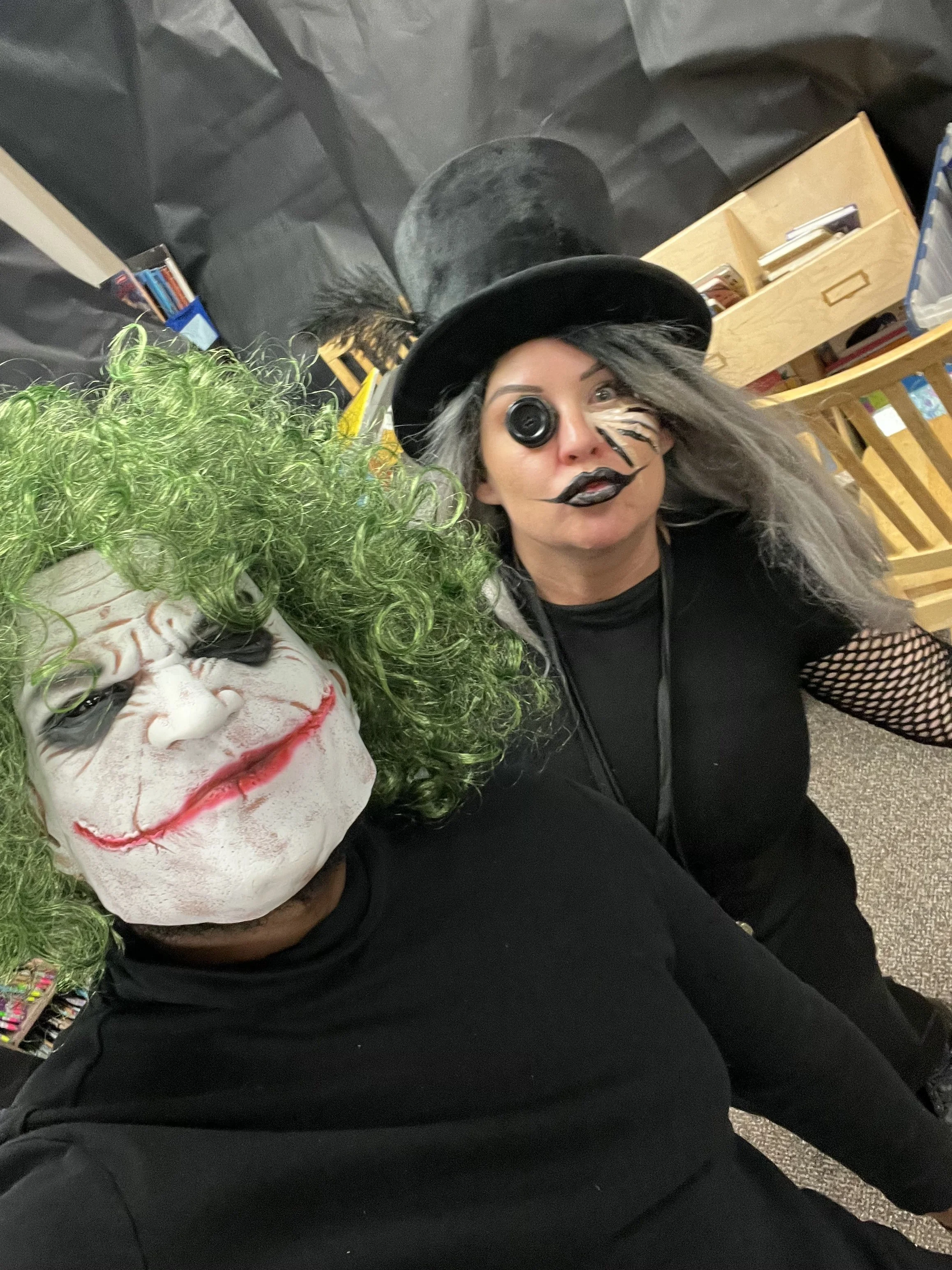 A person dressed as the Joker character from Batman is wearing a white face mask with green curly hair, red lipstick, and a painted smile. They are dressed in black and are seated next to a person with long gray hair, makeup resembling a clown with black lines around the eyes, lips, and face paint. The person with gray hair is wearing a black witch's hat and black clothing with fishnet sleeve details. They are in an indoor setting, possibly a store or an event space, with shelves and boxes in the background.