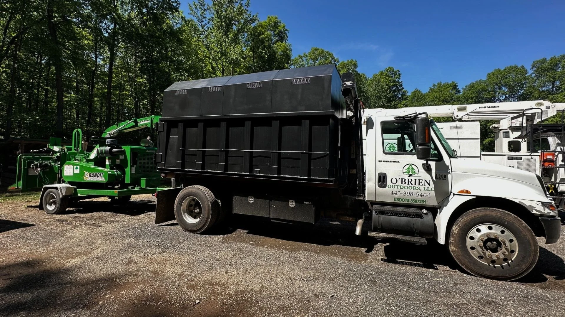 A white utility truck with a black enclosed bed and a crane, parked outdoors on gravel, with a green wood chipper attachment labeled 'Bandit' connected to it. The background features a wooded area with tall green trees and a clear blue sky.