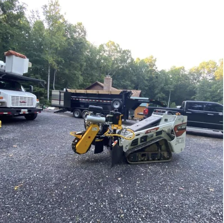 A black and yellow robotic construction or landscaping machine on asphalt, with a trailer, truck, and black pickup truck parked nearby, and trees and a house in the background.