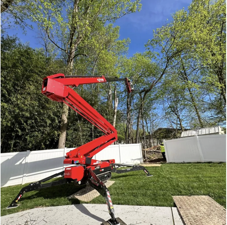A red aerial lift vehicle with extended arm and bucket, positioned on a grassy backyard with a white fence and tall trees under a blue sky.