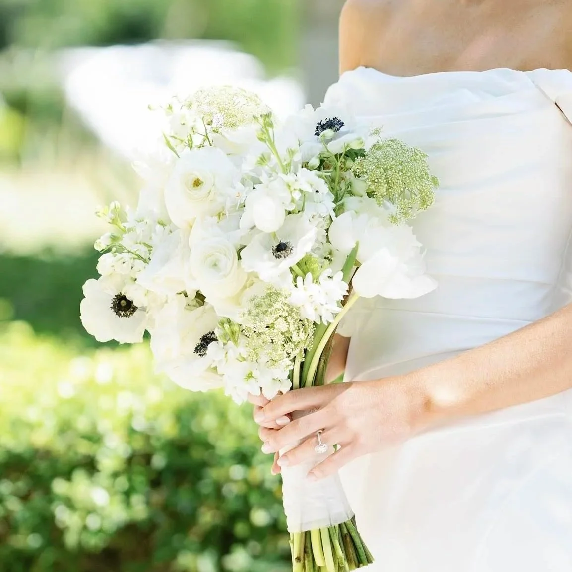 A bride holding an all white flower bouquet with a wedding dress and engagement ring, outdoors with green foliage in the background.
