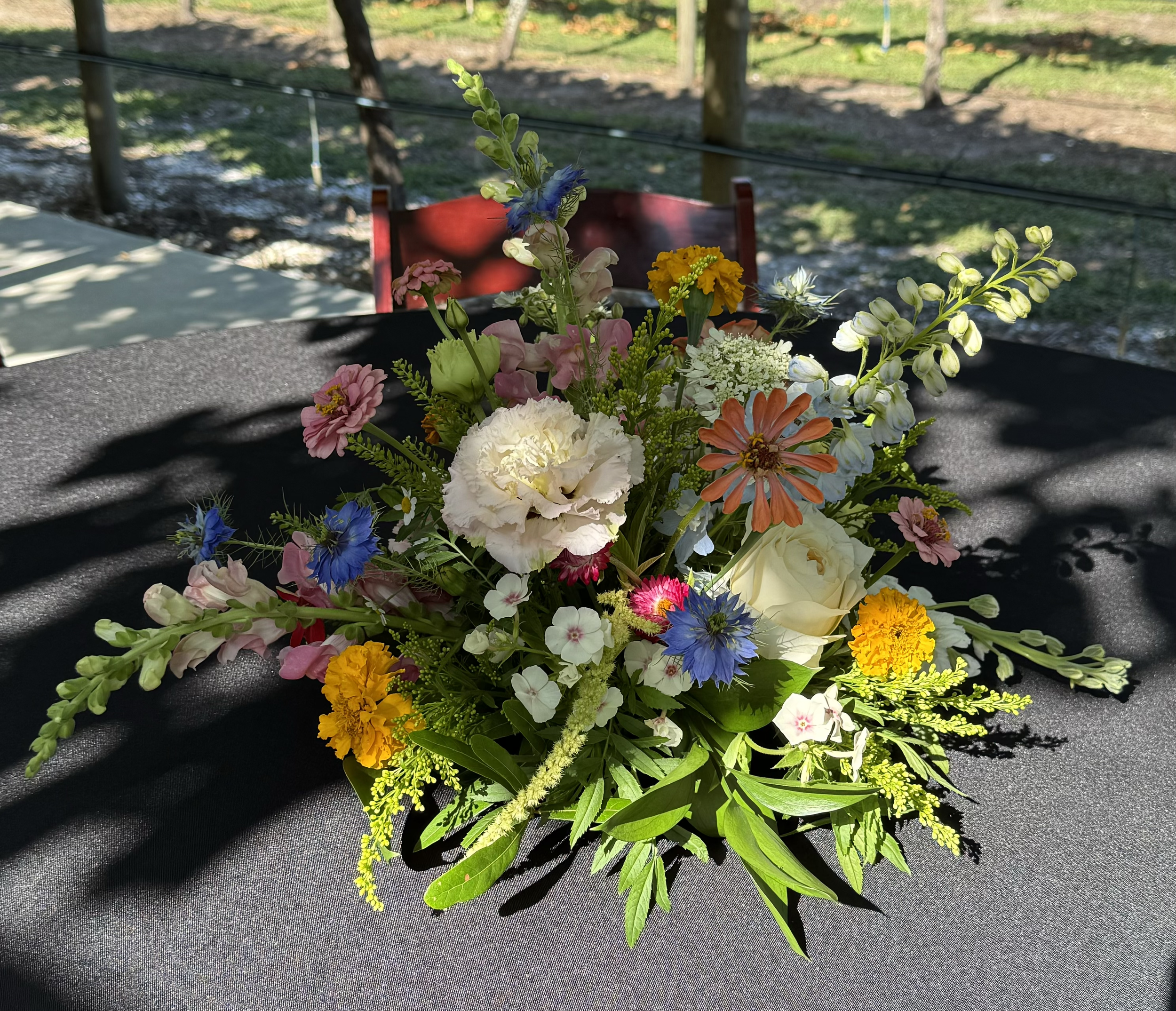 A centerpiece of mixed flowers including pink, white, yellow, orange, and blue blooms on a black table with an outdoor background of trees and grass.