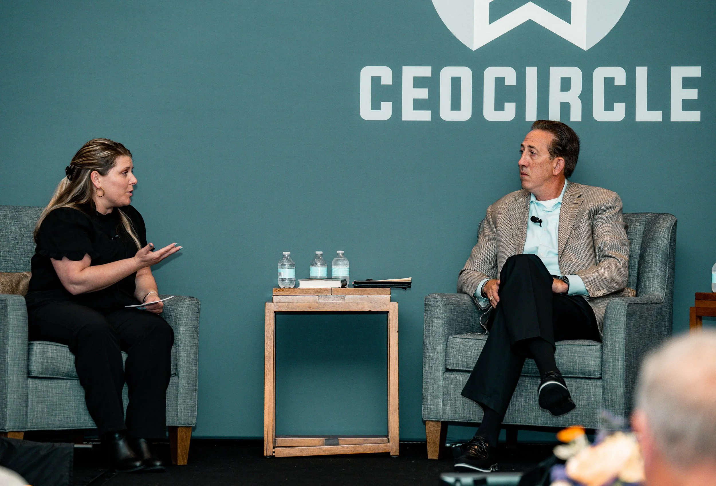 Two individuals seated in a discussion, with a teal wall behind them and the word 'CEOCIRCLE' visible. They have water bottles, notebooks, and microphones in front of them.
