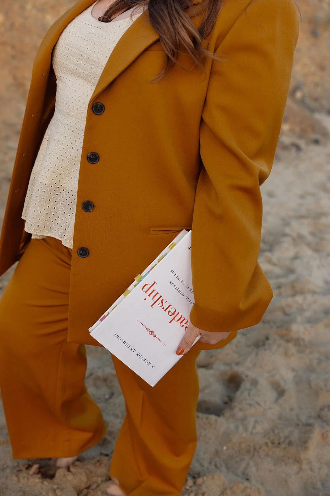 A person wearing a mustard yellow suit and white eyelet top, holding a white book with orange and black text, standing on a sandy surface.