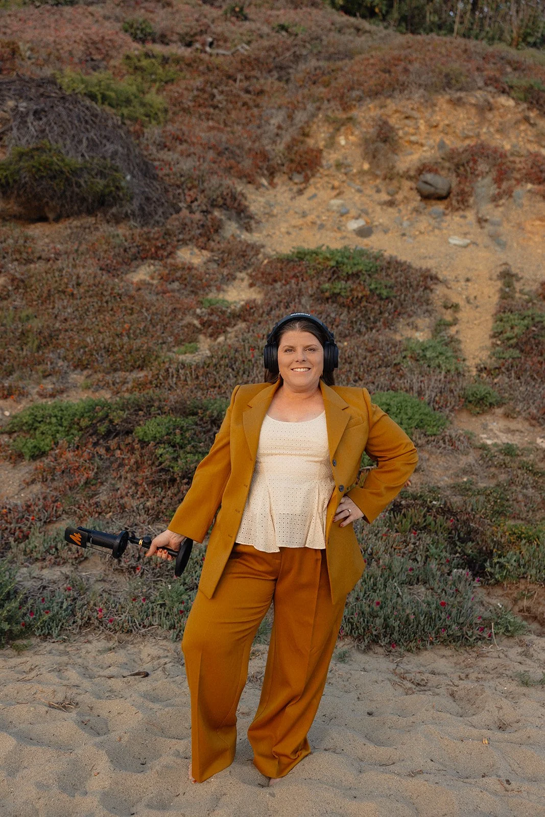 A woman in a mustard-colored suit and beige blouse, standing on a sandy beach with her hand on her hip, wearing black headphones, holding a metal detector in her right hand, and smiling at the camera, with low, rocky hills and sparse vegetation in the background.
