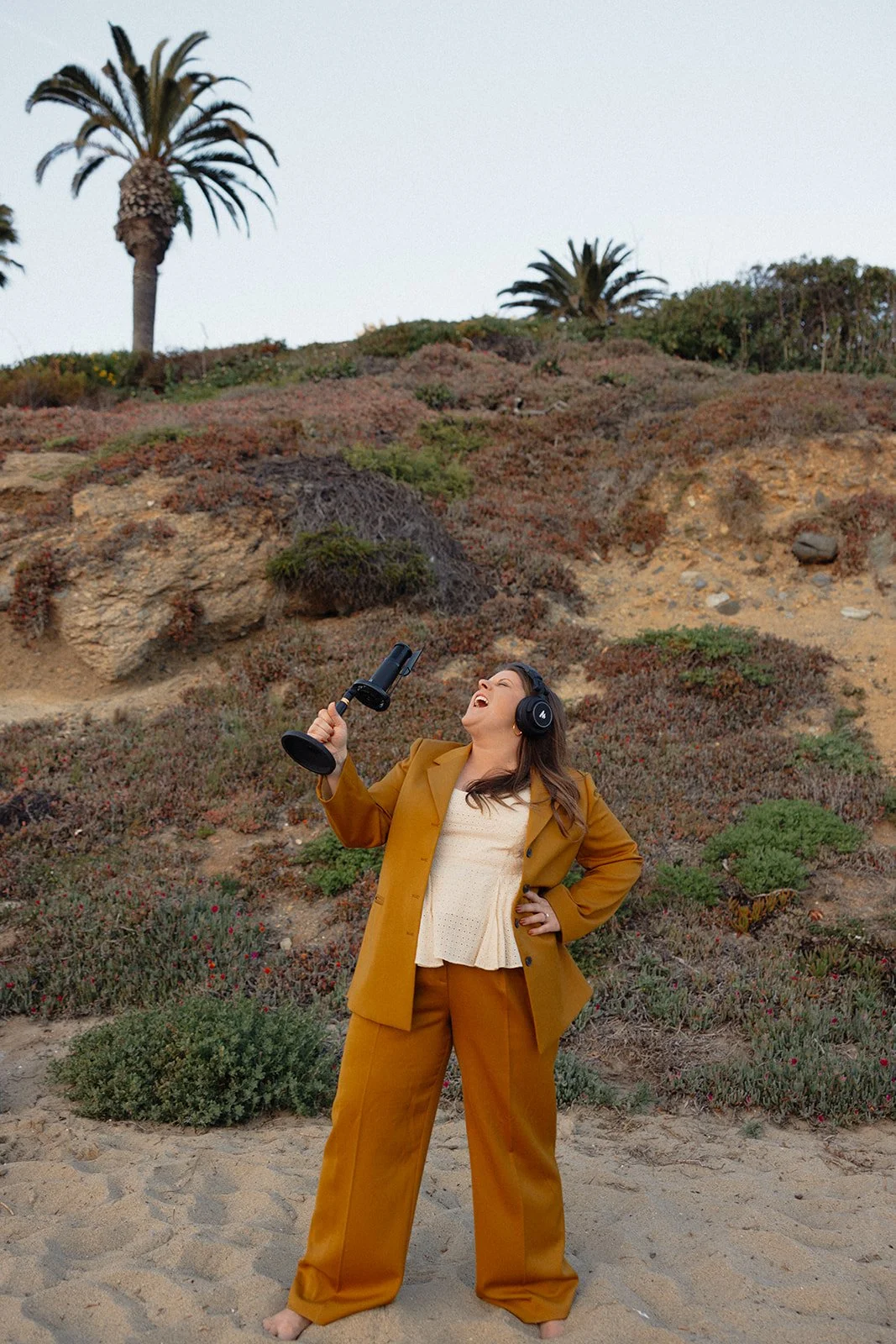 A woman standing on a sandy beach, wearing a mustard-colored suit and headphones, holding a microphone and singing or speaking, with palm trees and coastal vegetation in the background.