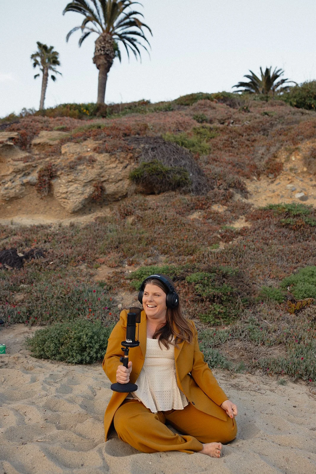 Woman sitting on the sandy beach, holding a microphone and wearing headphones, with a hillside covered in greenery, rocks, and desert plants, and palm trees in the background.
