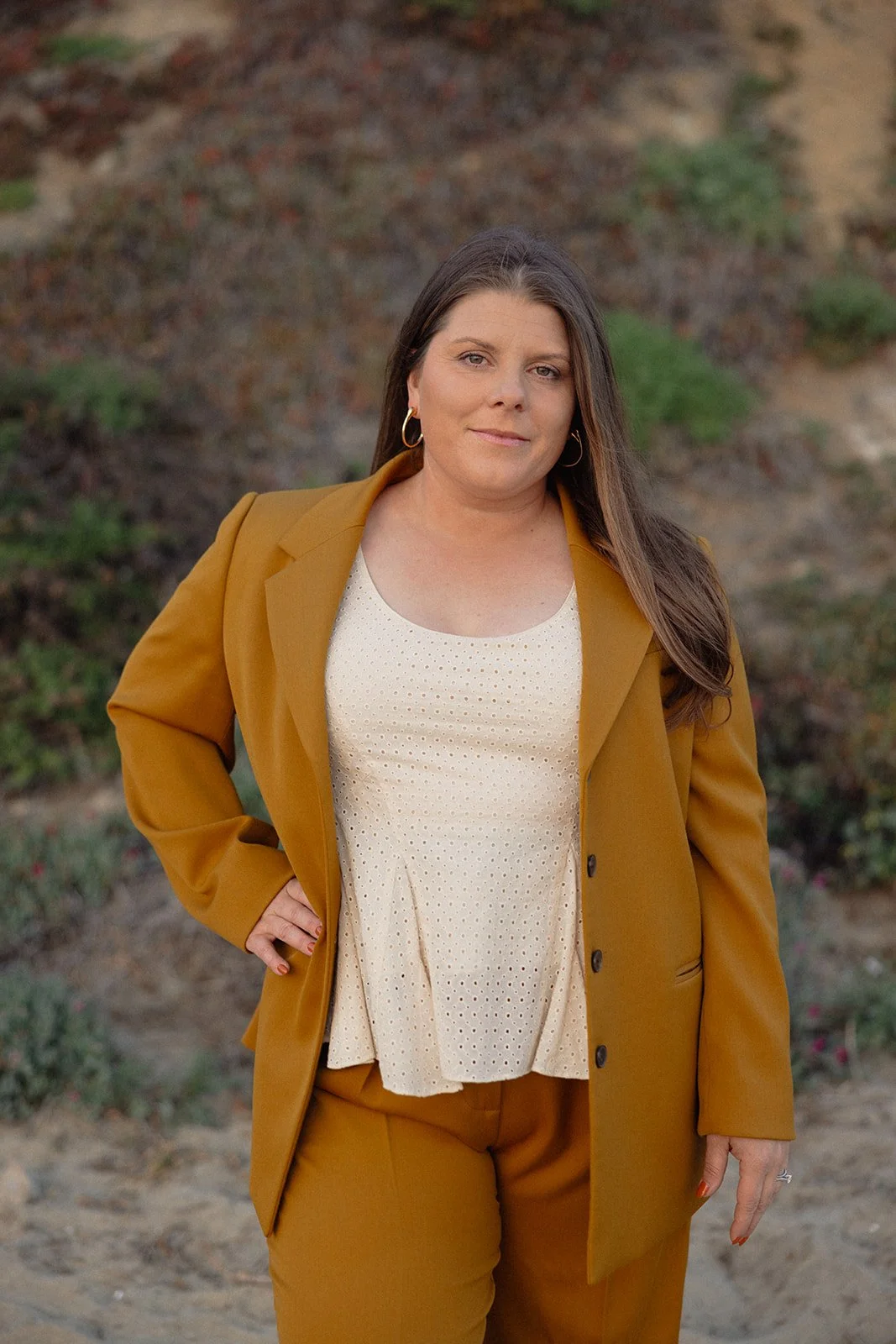 A woman with long brown hair wearing gold hoop earrings, a mustard yellow blazer, and matching pants, standing outdoors in front of a hillside with greenery and soil.