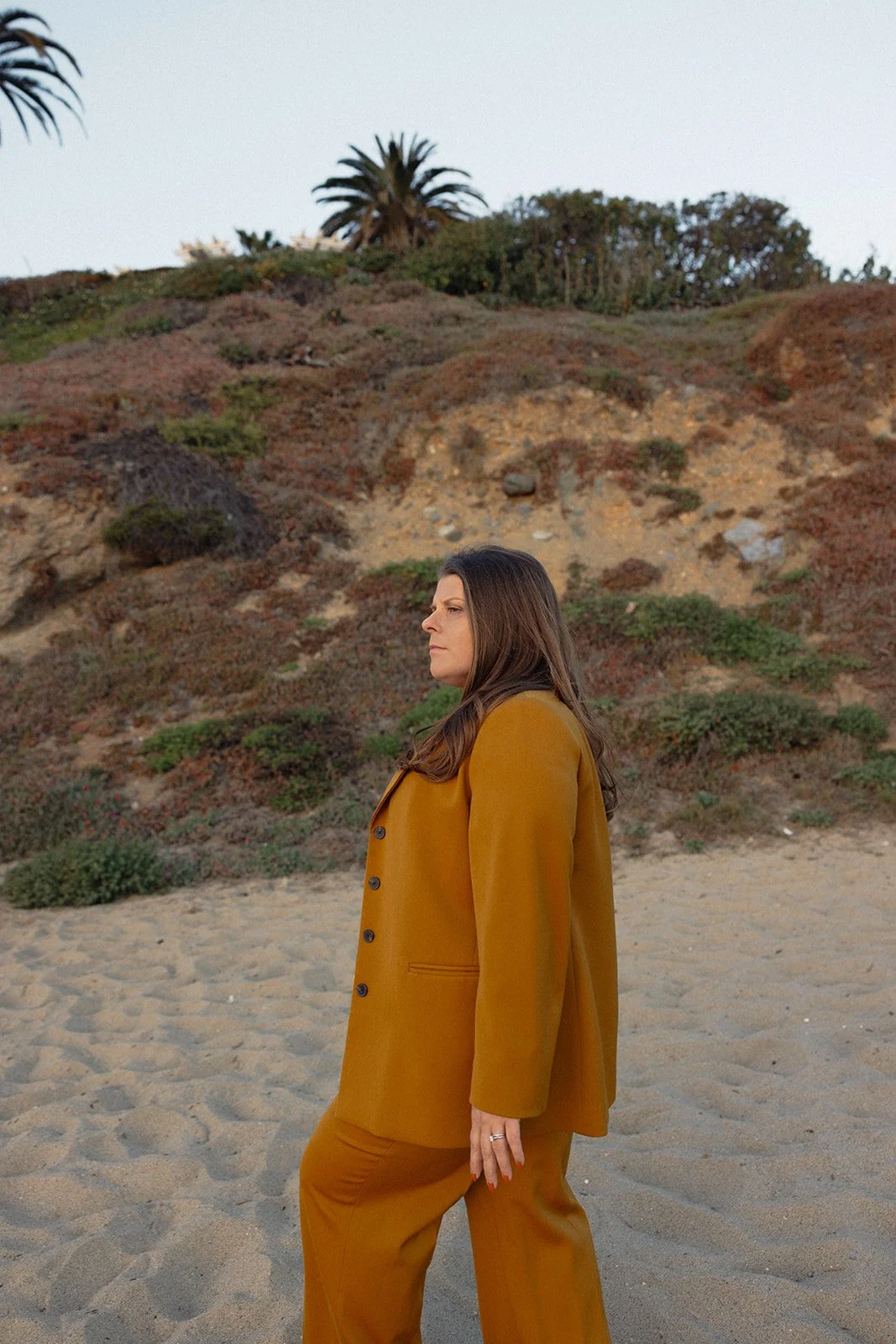 A woman in a mustard yellow suit standing on a sandy beach with a hillside covered in plants and trees in the background.