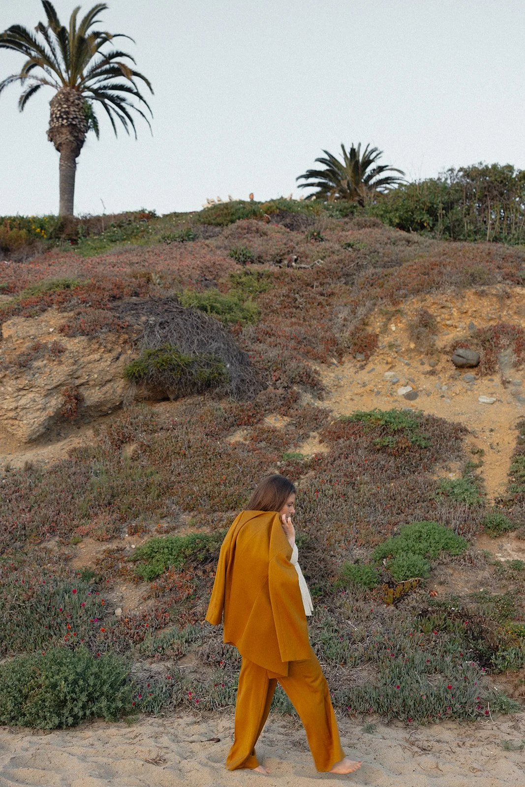 A woman walking barefoot on a sandy beach with dry bushes and rocks on a hill behind her, and palm trees at the top of the hill.